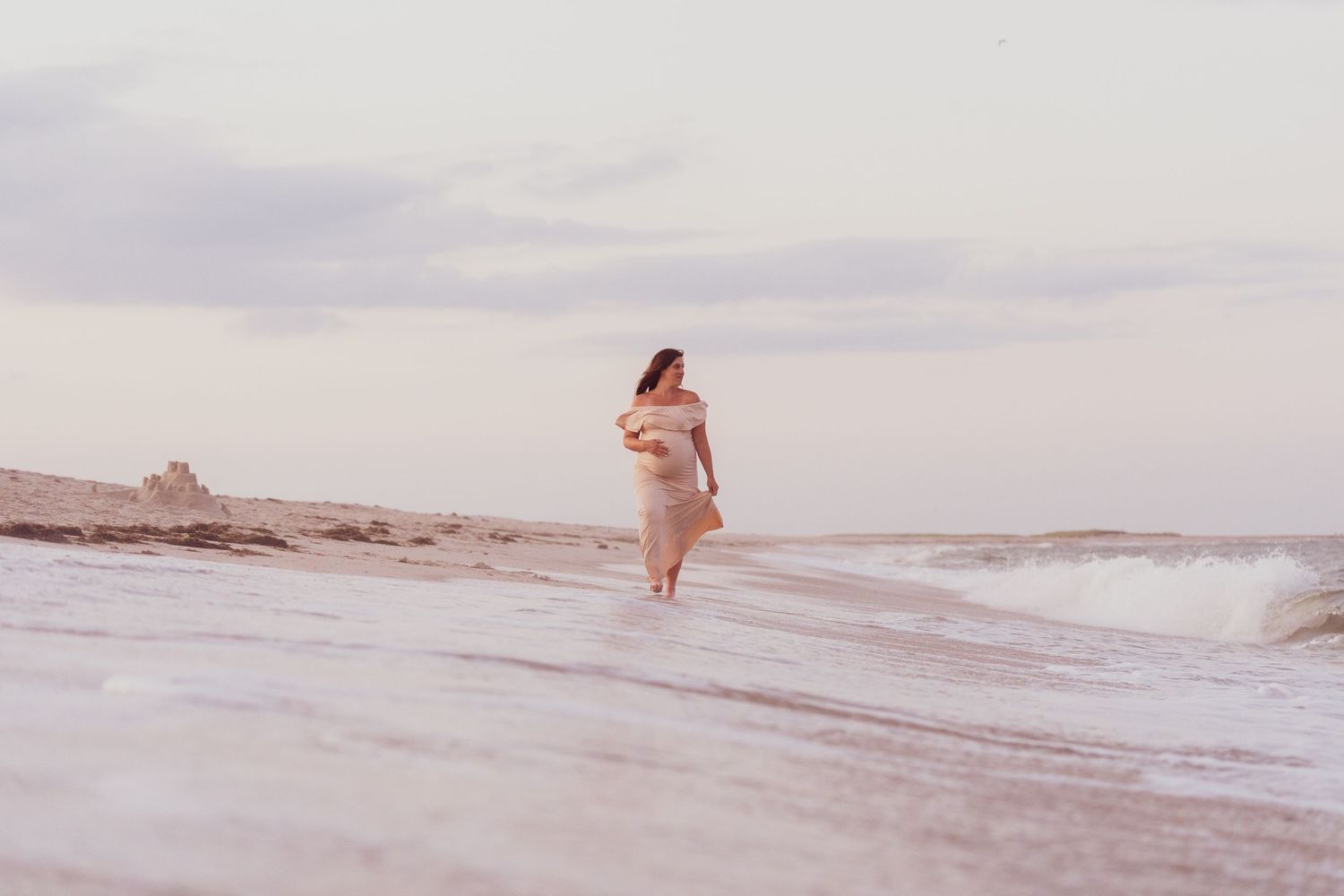 A person in a flowing dress walks along a misty beach shoreline at sunset with waves rolling in.