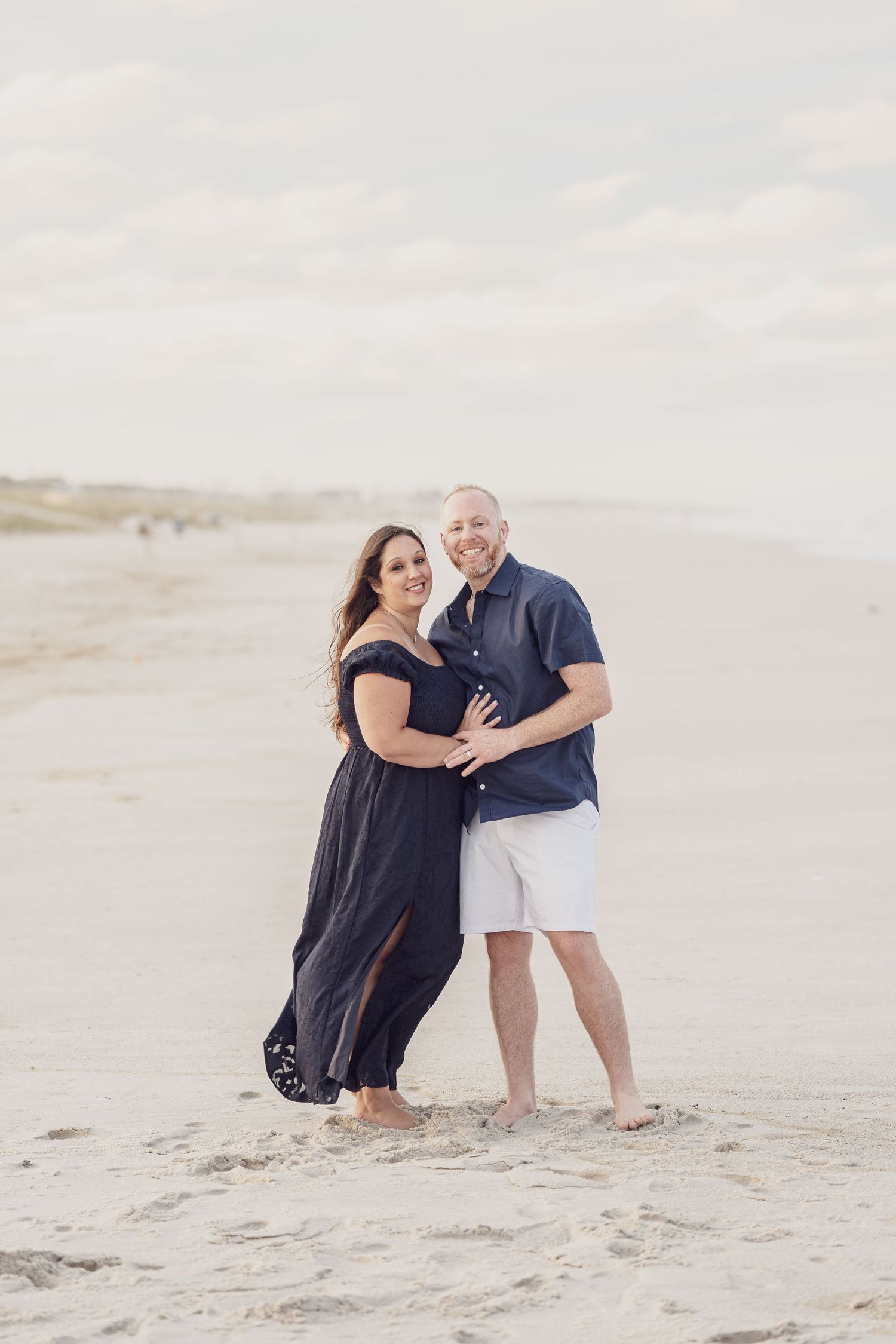 A couple in dark clothing shares a tender moment on sand dunes during a beach photoshoot.