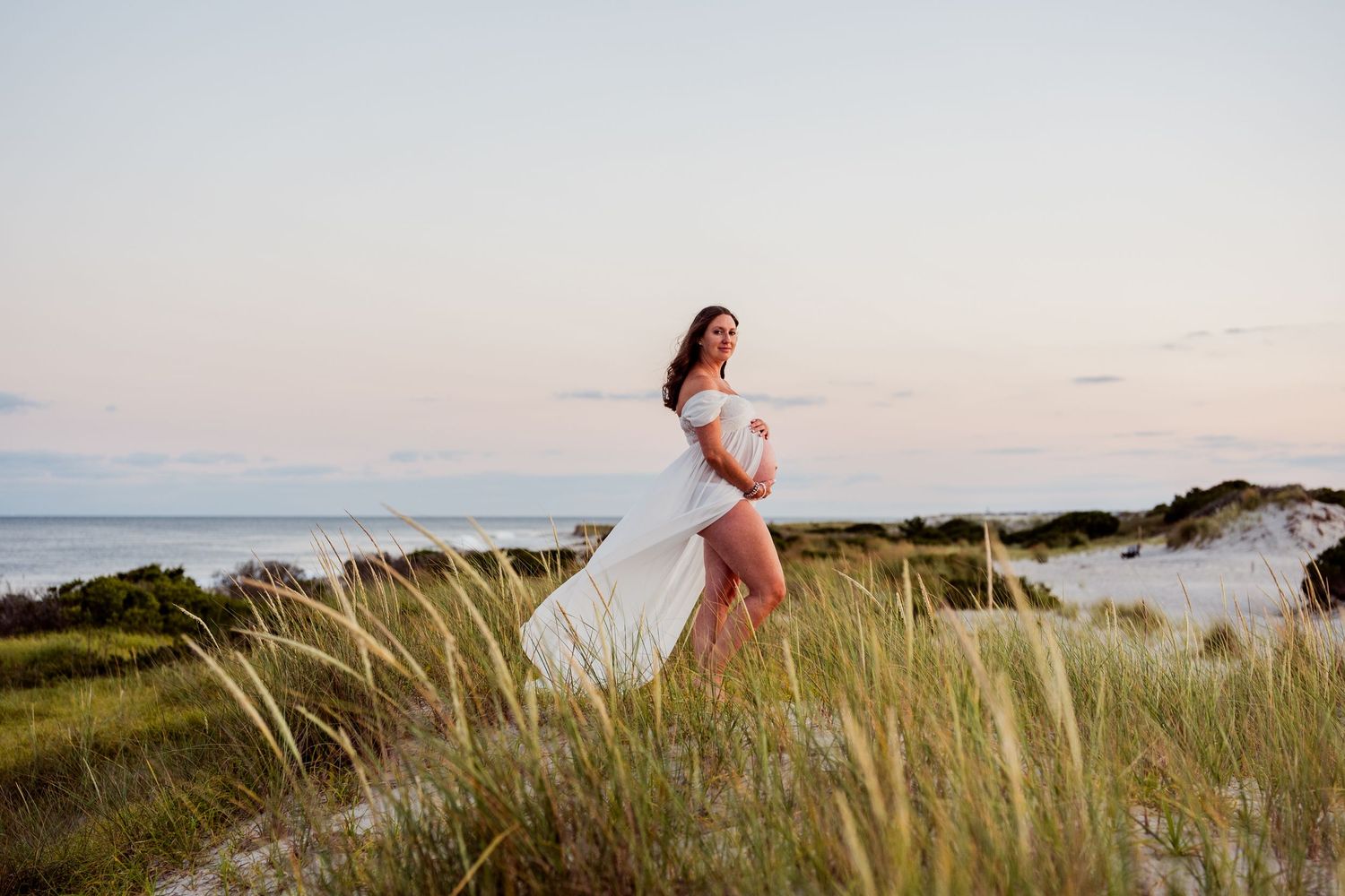 Person in flowing dress poses on coastal dunes during sunset.
