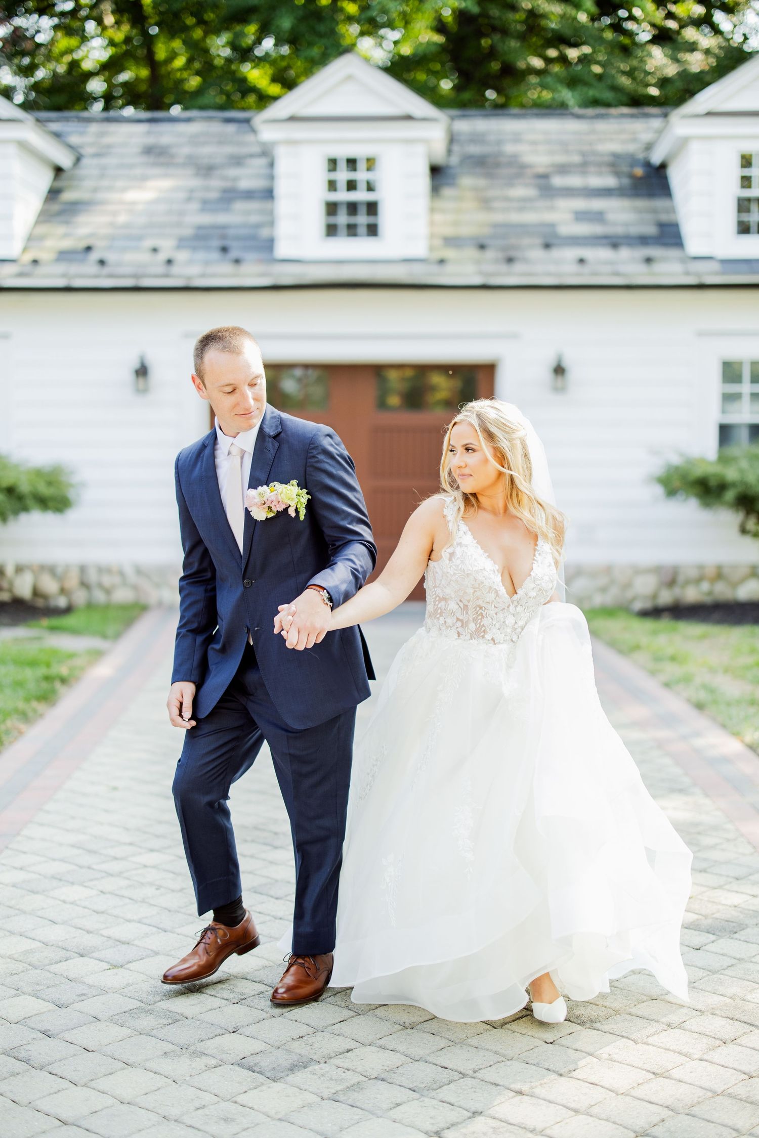 Couple in wedding attire playfully walking on brick pathway by white building.