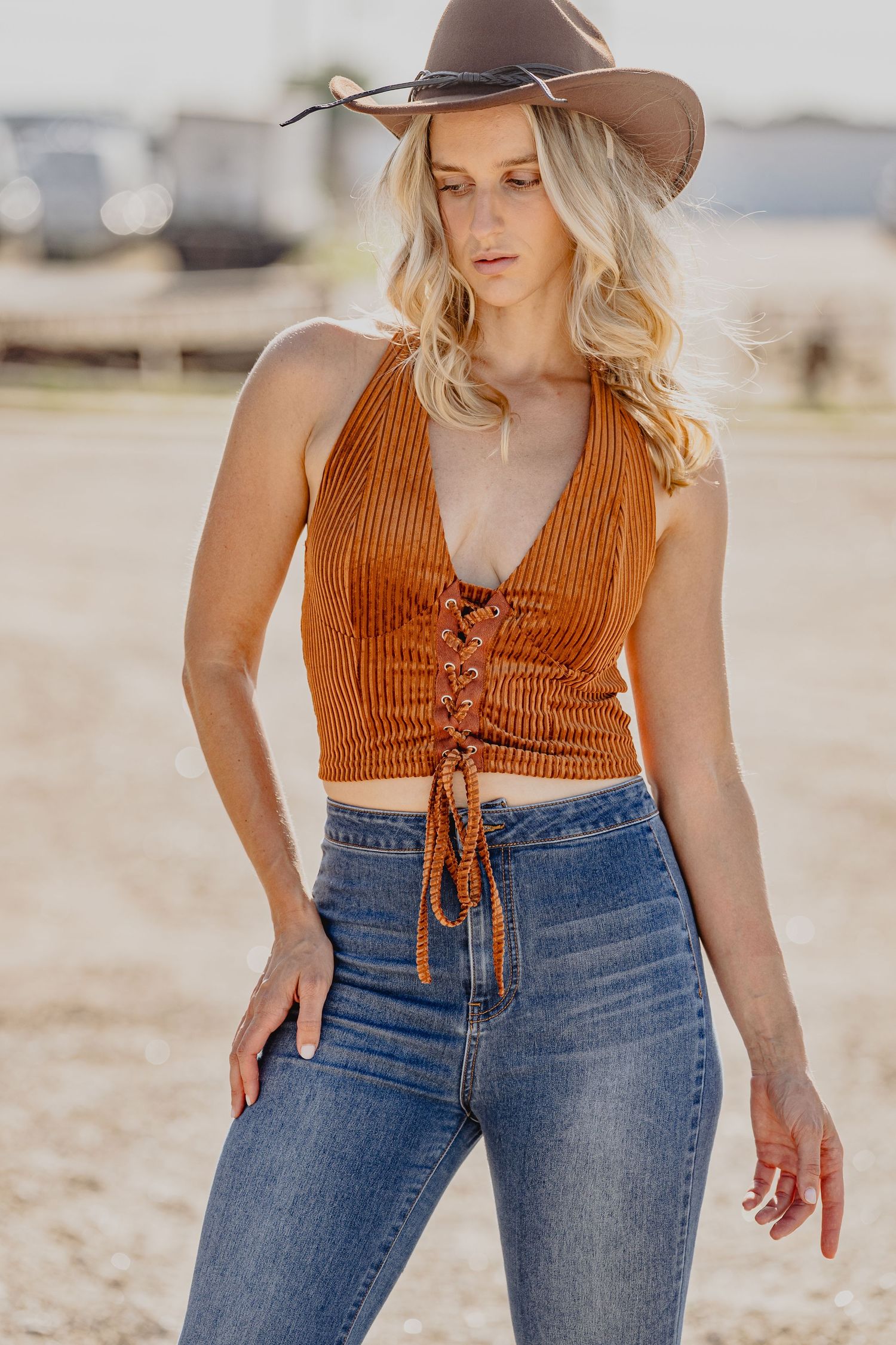 Blonde woman in rust orange lace-up halter crop top, jeans, and brown cowboy hat posing outdoors.