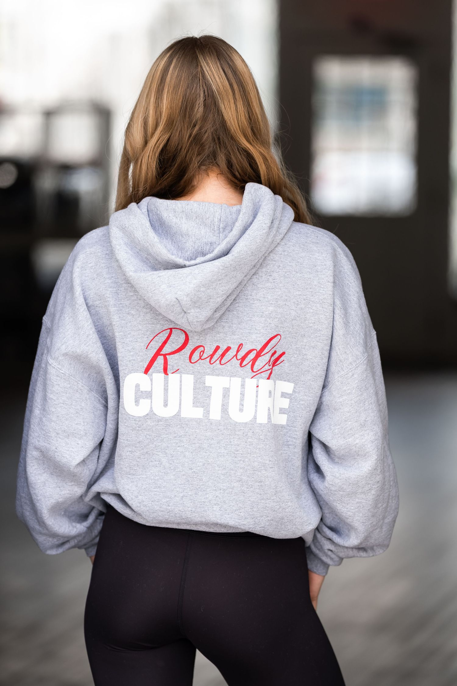 Woman wearing a gray Rowdy Culture hoodie with black leggings, shown from behind.