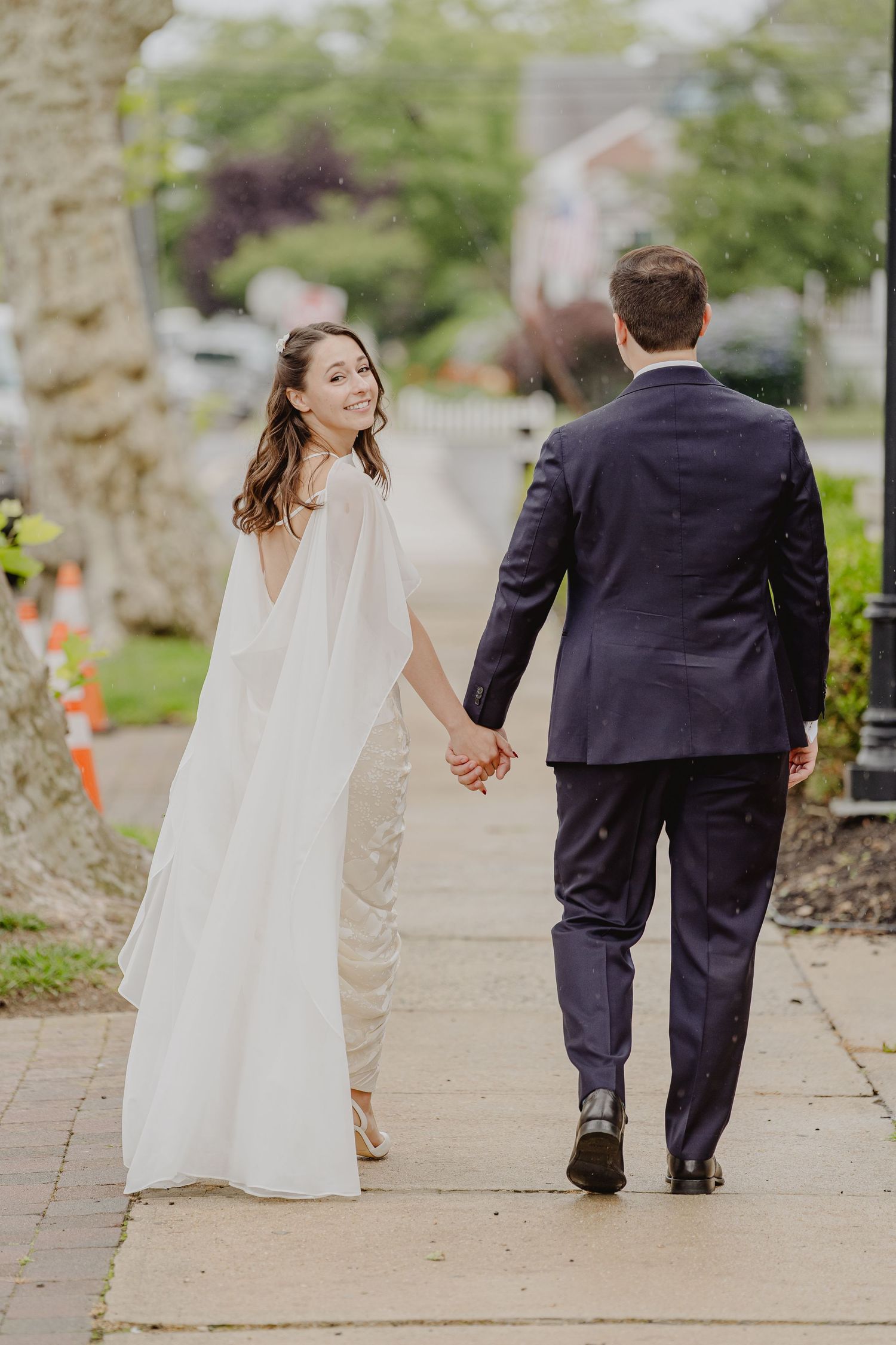 Bride in white cape gown and groom in navy suit walk hand-in-hand on a sidewalk, bride smiling back at camera.