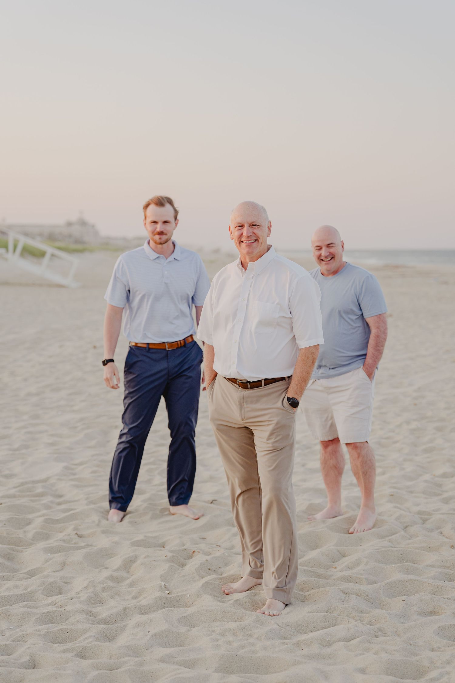 Three men posing barefoot on sandy beach at sunset, wearing casual summer attire.