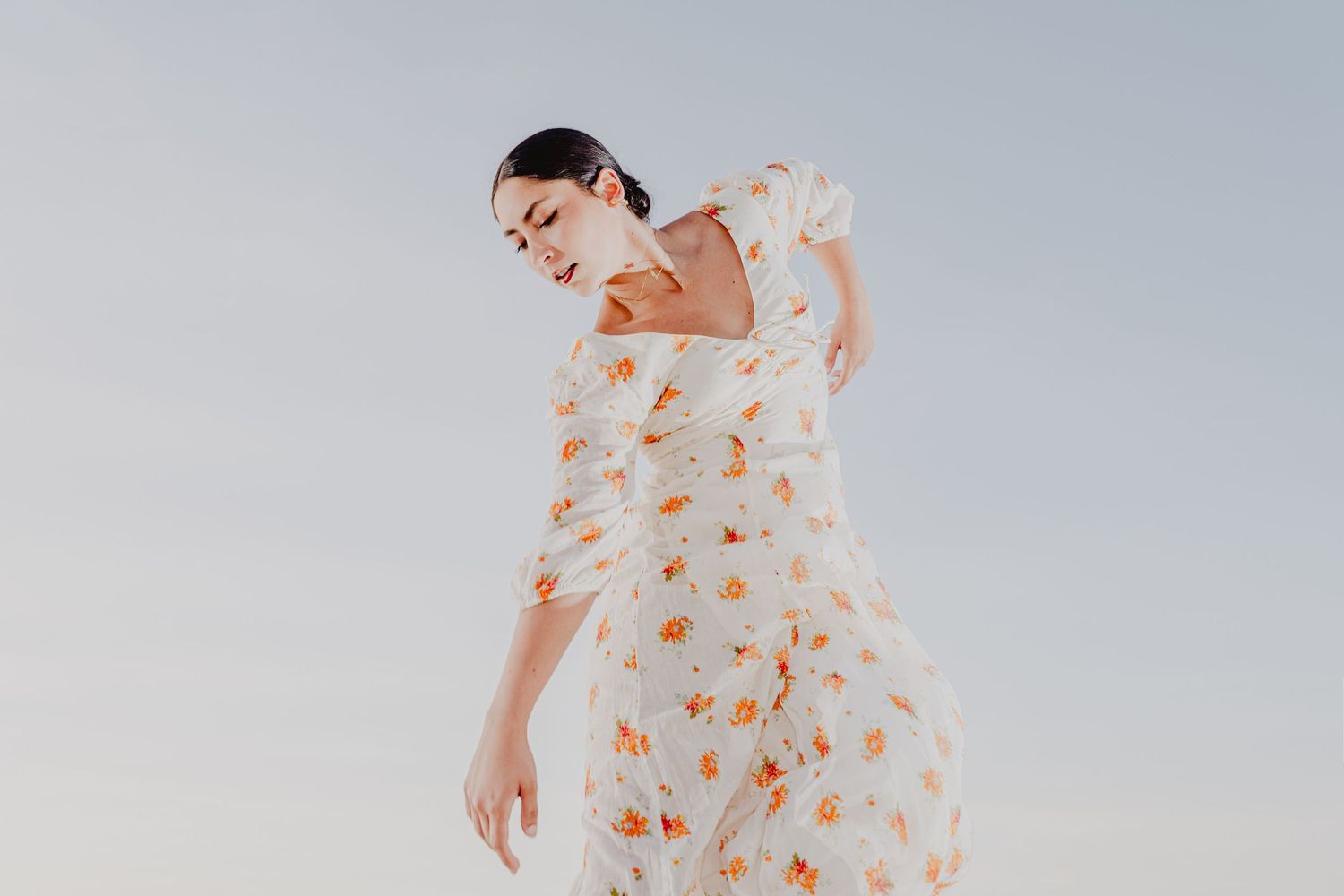 Woman in flowing white floral dress with orange blooms poses gracefully against a soft blue sky.