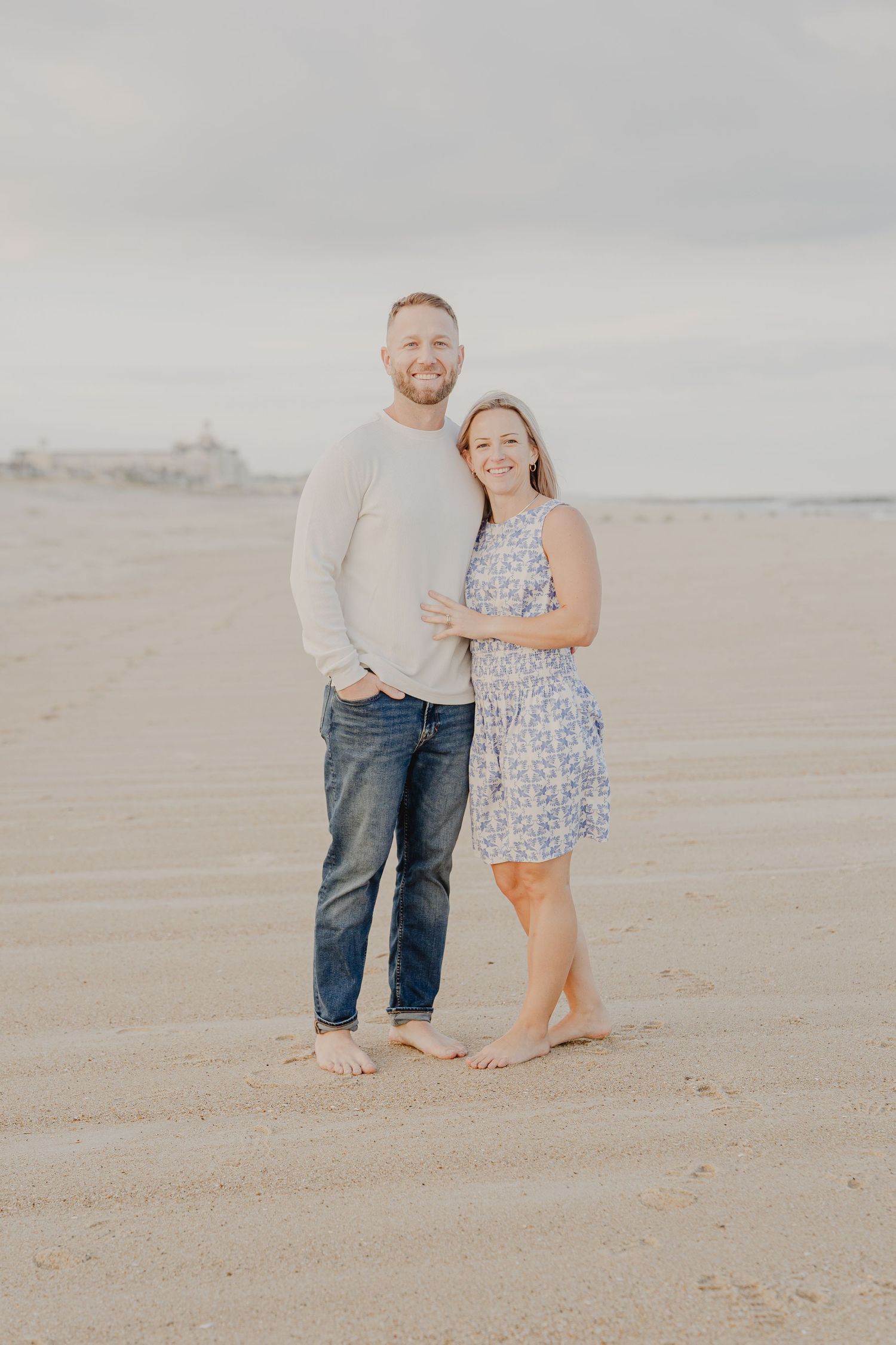 Smiling barefoot couple embraces on sandy beach, man in white sweater and jeans, woman in blue floral dress.