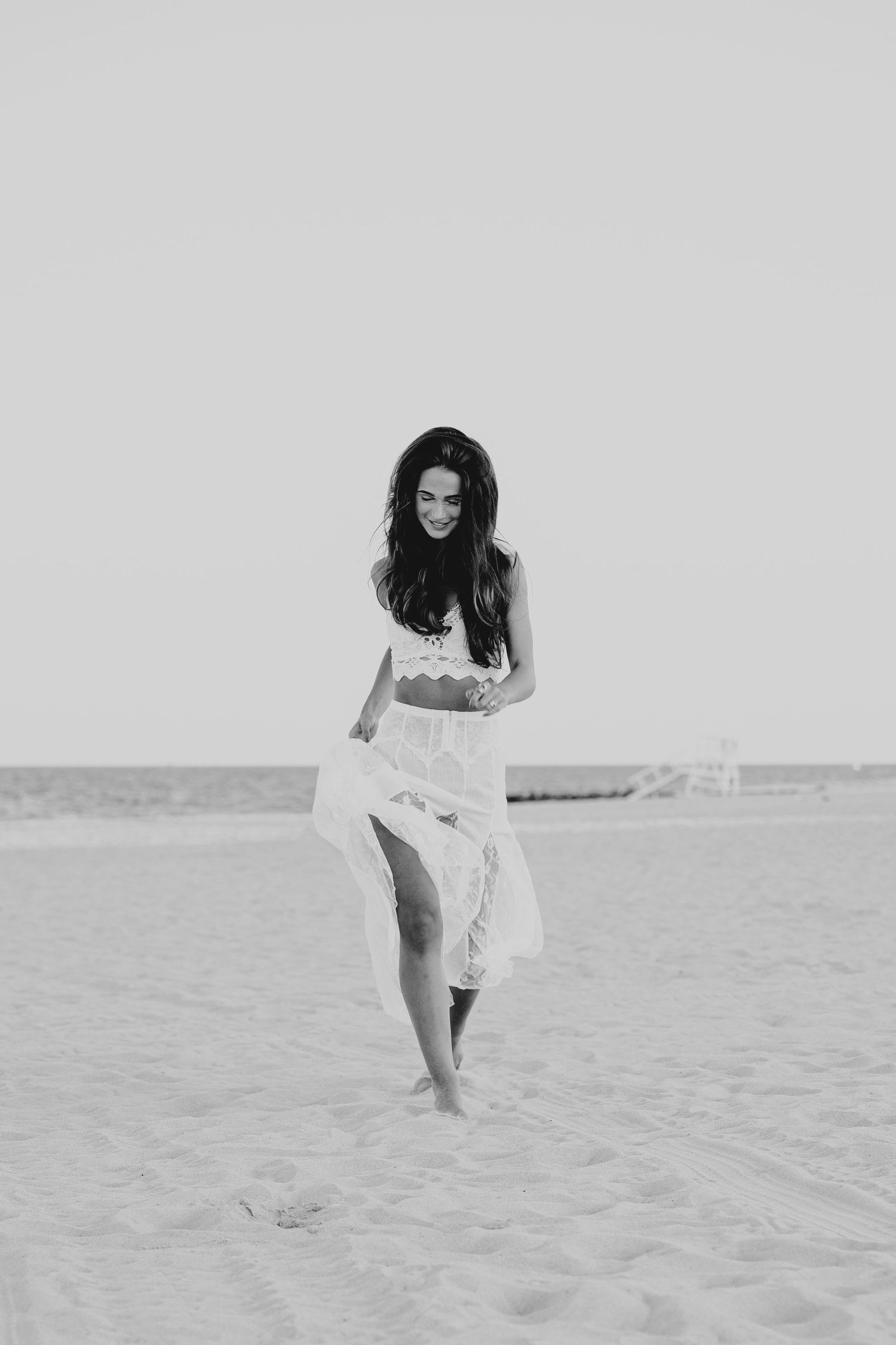 A smiling woman in a white crop top and flowing skirt walks barefoot on a sandy beach in black and white photo.