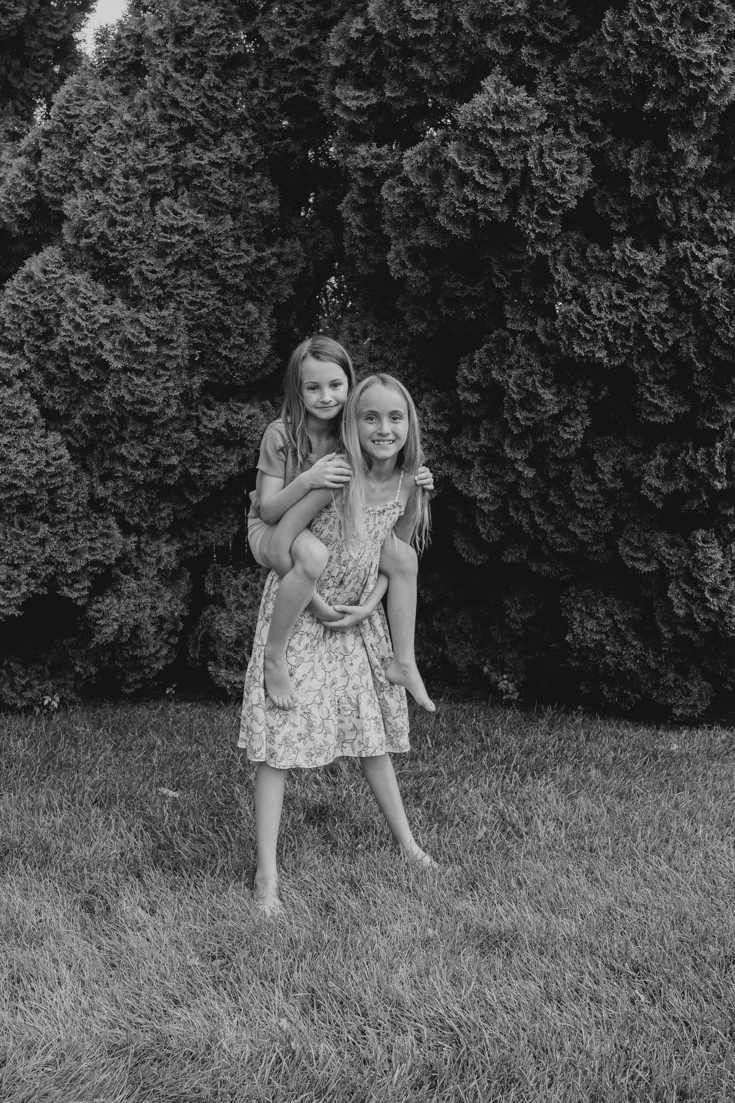 Two young girls in floral dresses pose together outdoors, one giving the other a piggyback ride in front of evergreen trees.