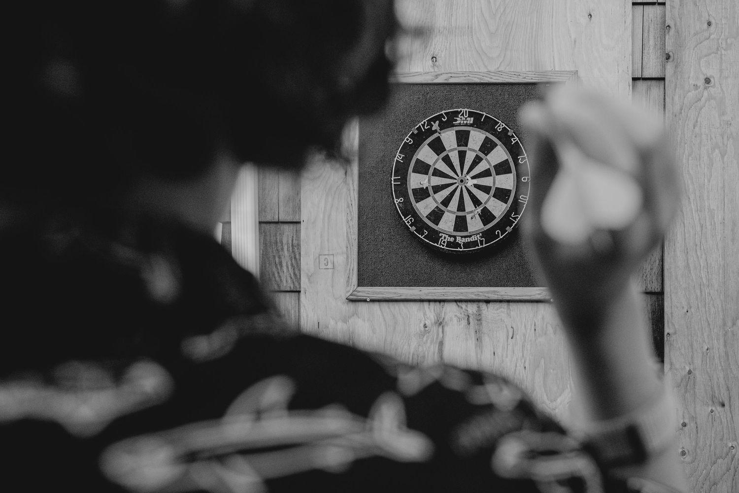 Black and white photo of a person throwing darts at a dartboard mounted on a wooden wall.