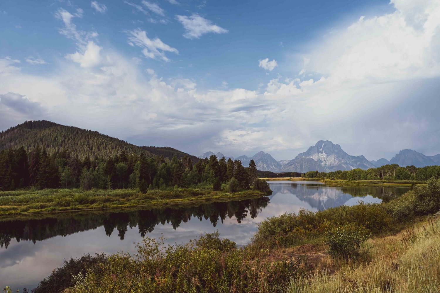 Panoramic view of Grand Teton National Park with mountains, lakes and meadows under blue skies with puffy clouds.
