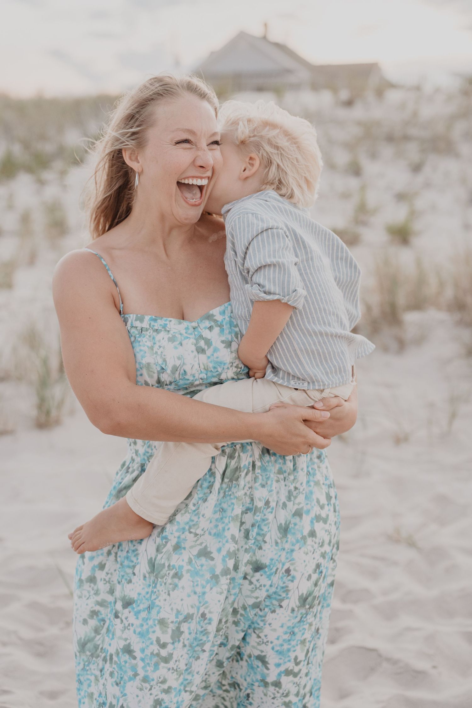 Mother and child sharing laughs and hugs on sandy beach in summer attire.