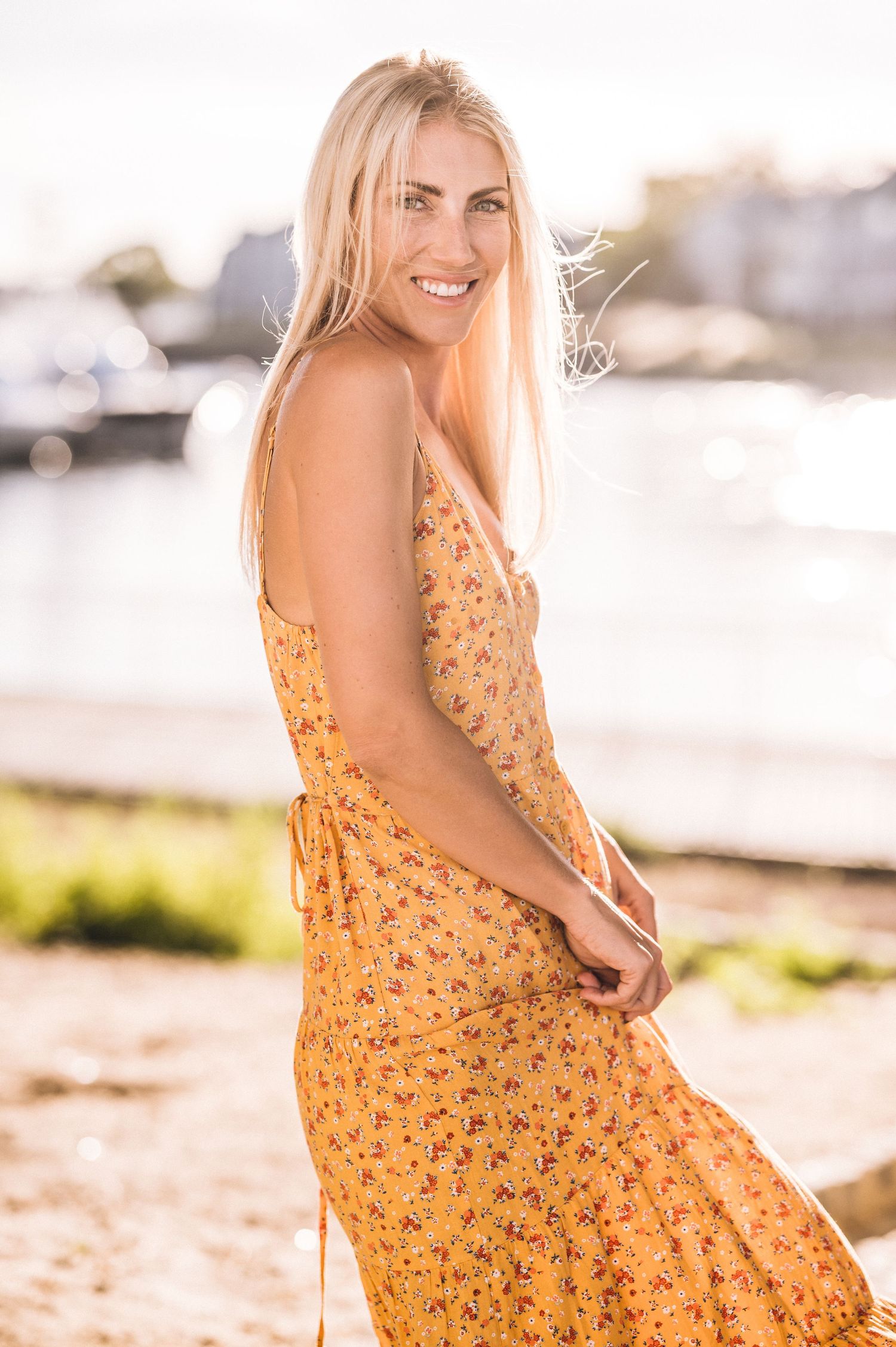 A woman in a yellow floral sundress sits on a sunny beach with boats in the background.
