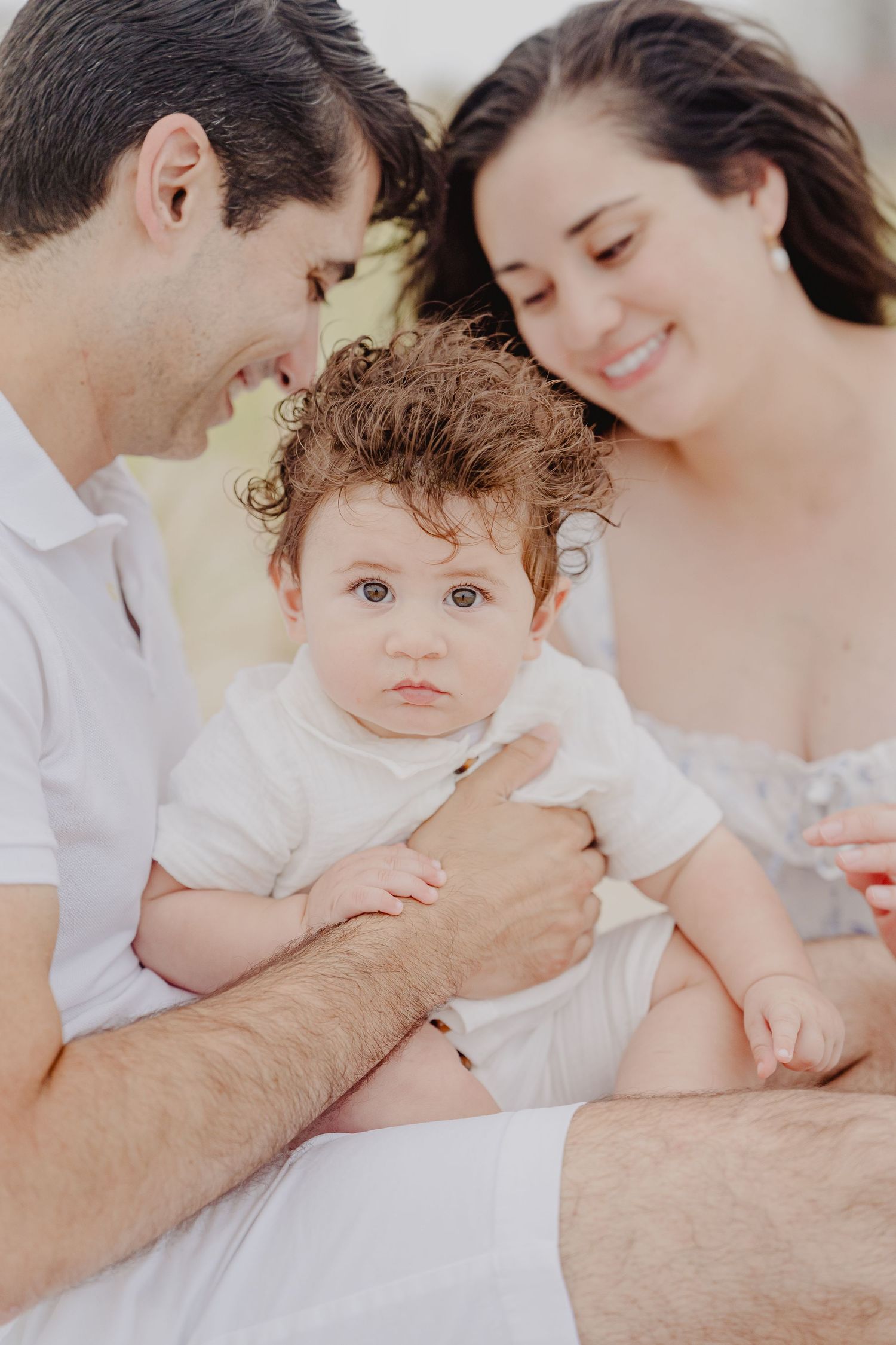 A happy young family with a curly-haired baby in white outdoors in soft natural light.