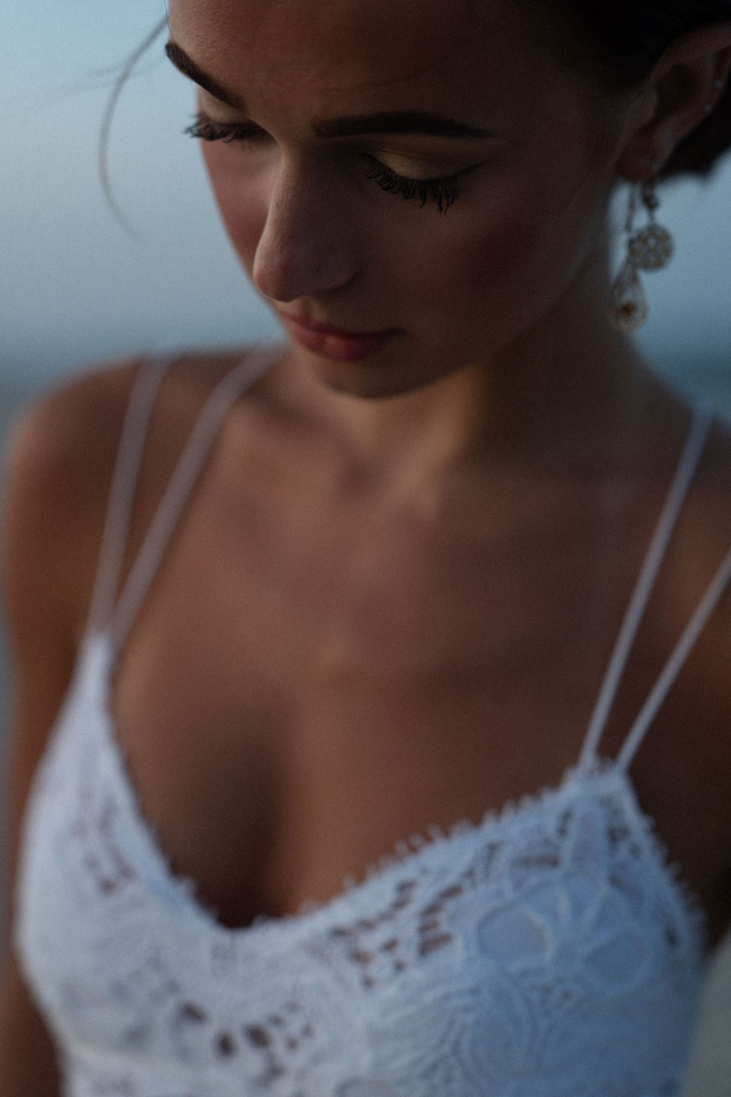 Young woman in white lace dress with elegant earrings, gazing downward in soft evening light.