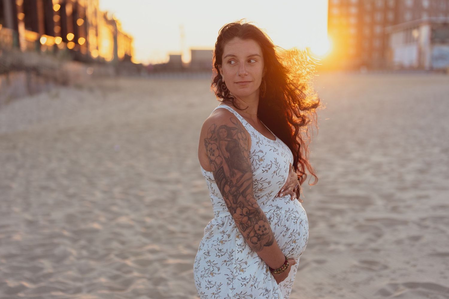 A pregnant woman in white lace dress poses on beach during sunset.