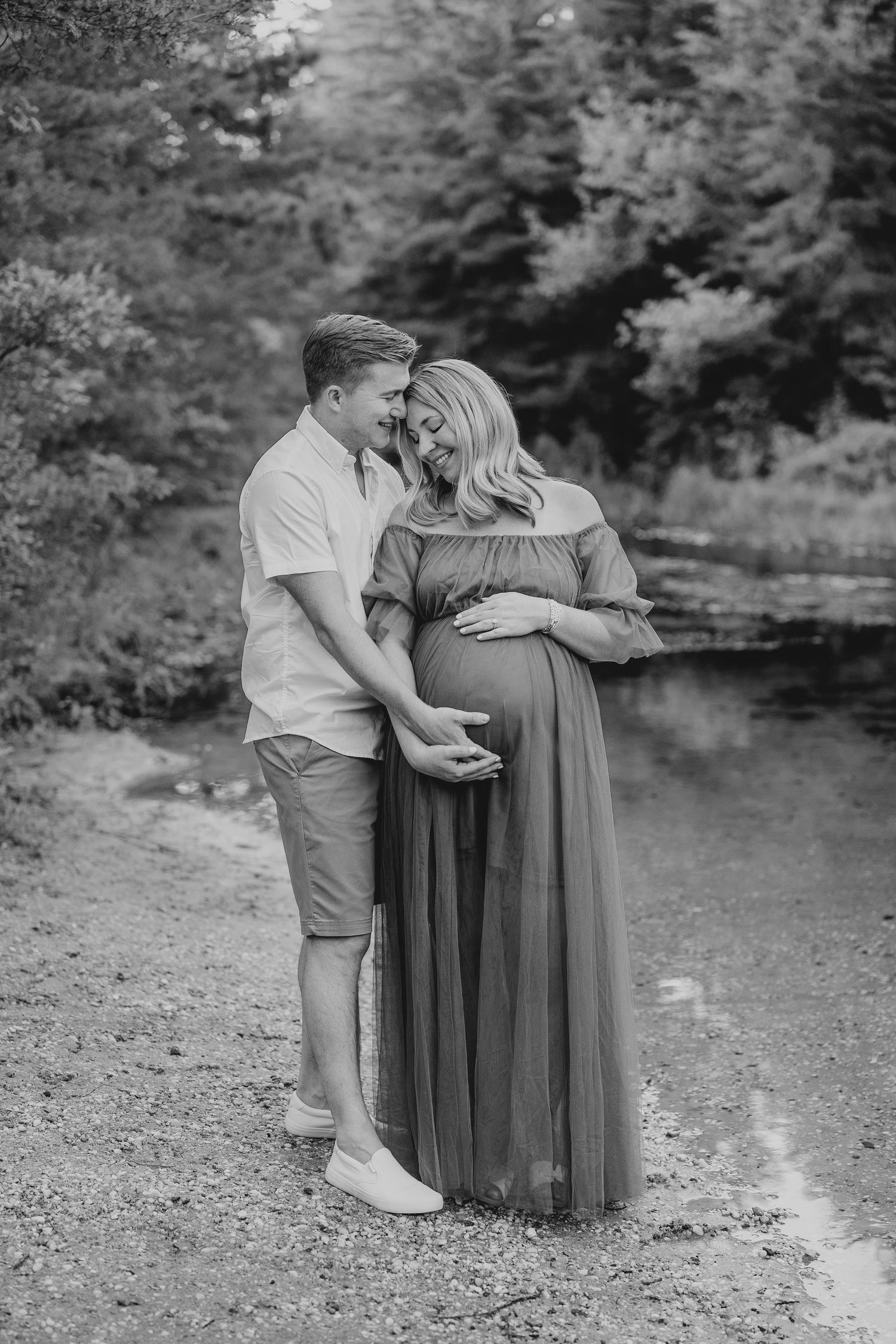 Black and white maternity photo by a pond with forest backdrop.