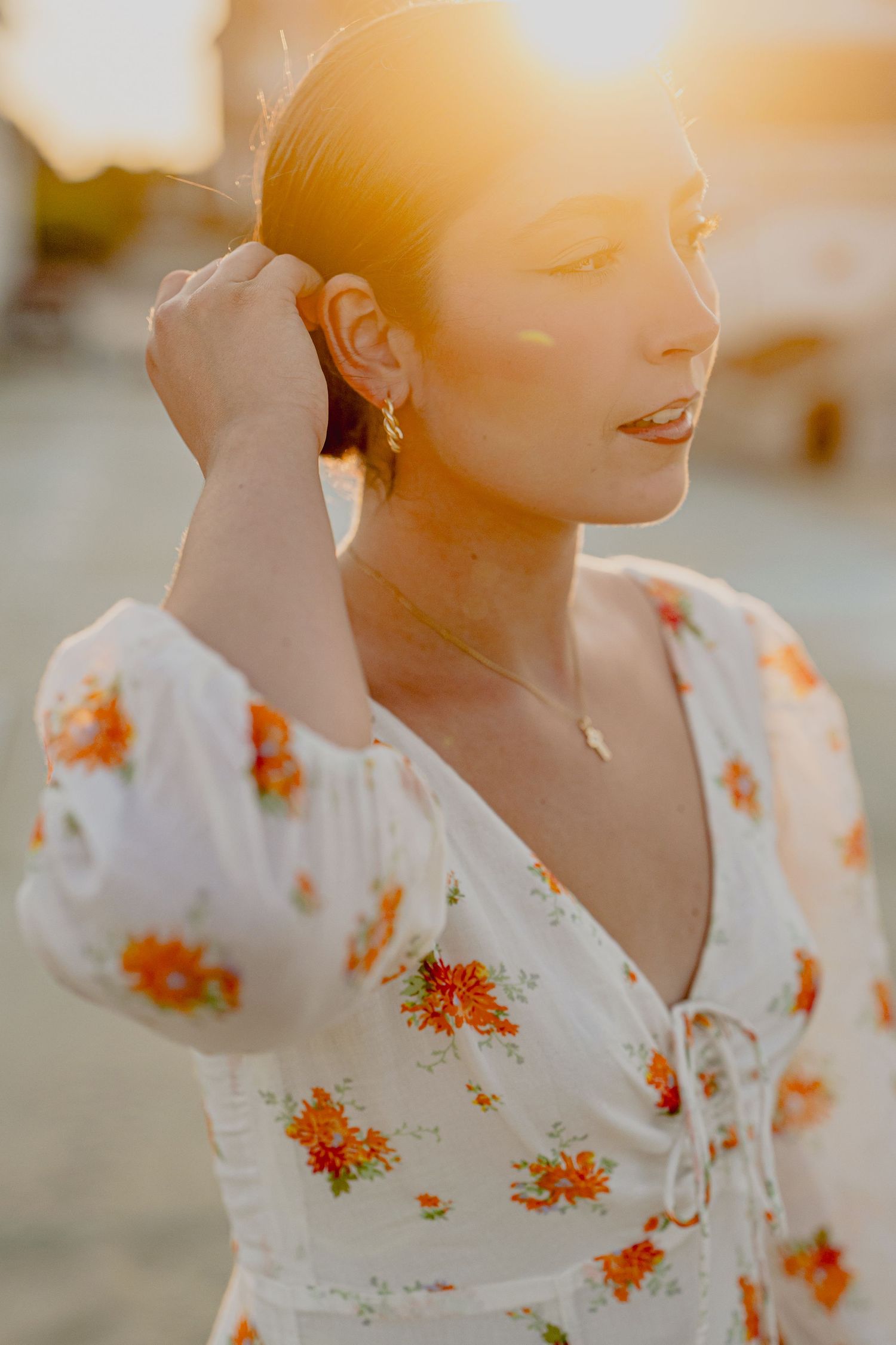 Woman in white floral dress with orange flowers poses in golden sunset light, touching her hair.