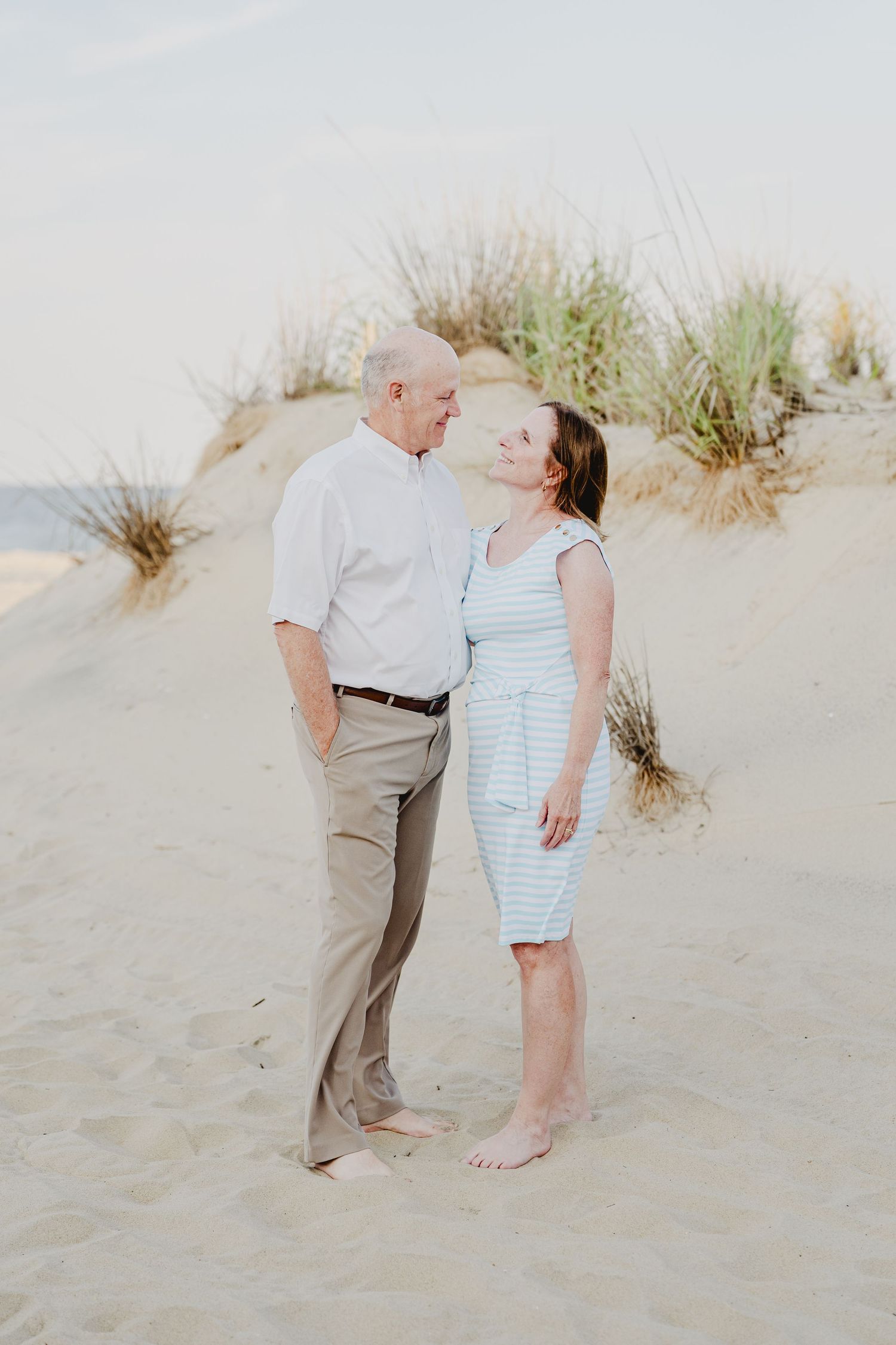 Couple sharing a romantic moment on sandy beach dunes, woman in striped dress and man in white shirt.