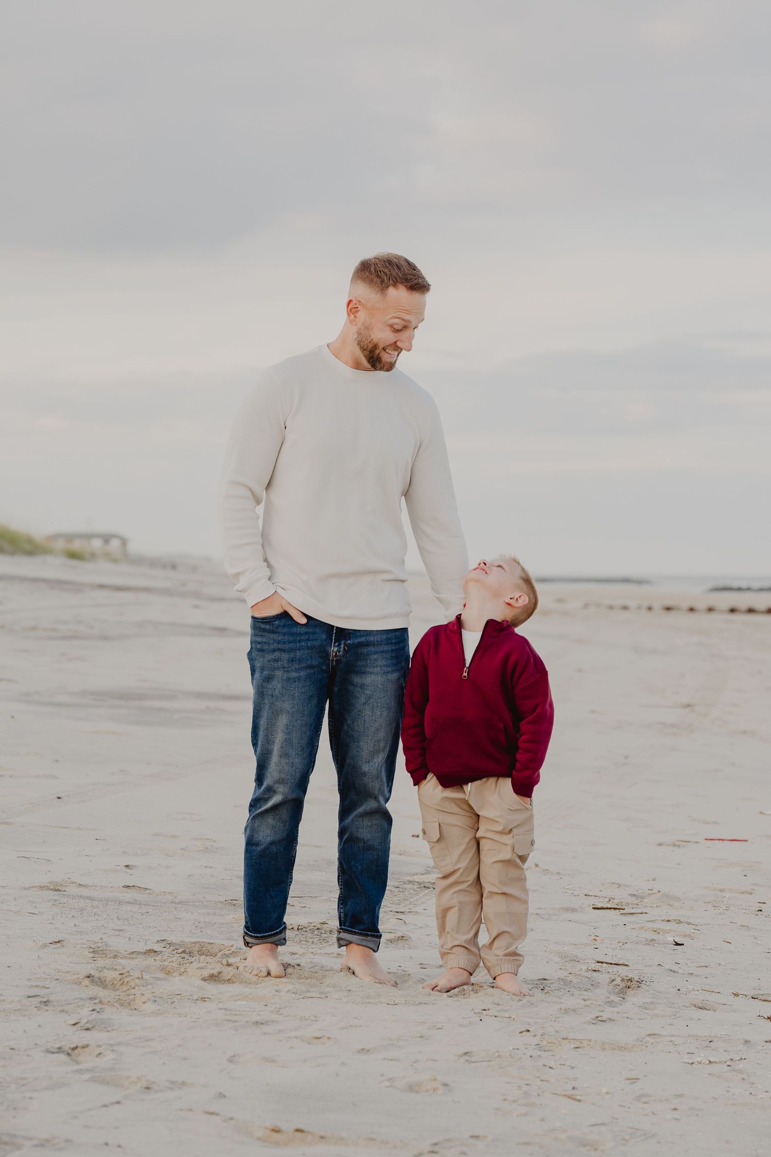 A father and young son share a joyful moment on a sandy beach, both barefoot and smiling at each other.