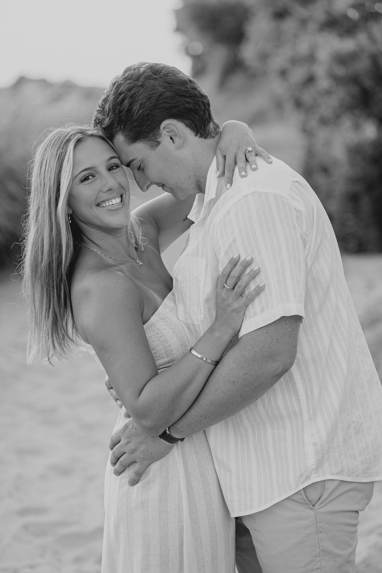 Black and white photo of a smiling couple embracing on a beach, woman in sundress, man in linen shirt.