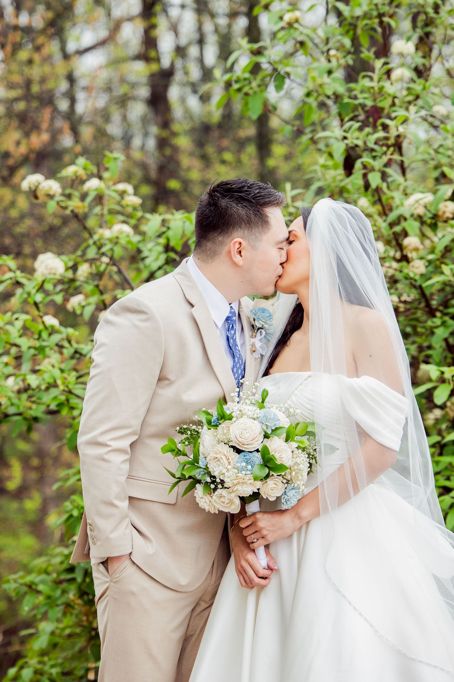 Romantic wedding couple shares a kiss in a garden setting with white florals and a tan suit.