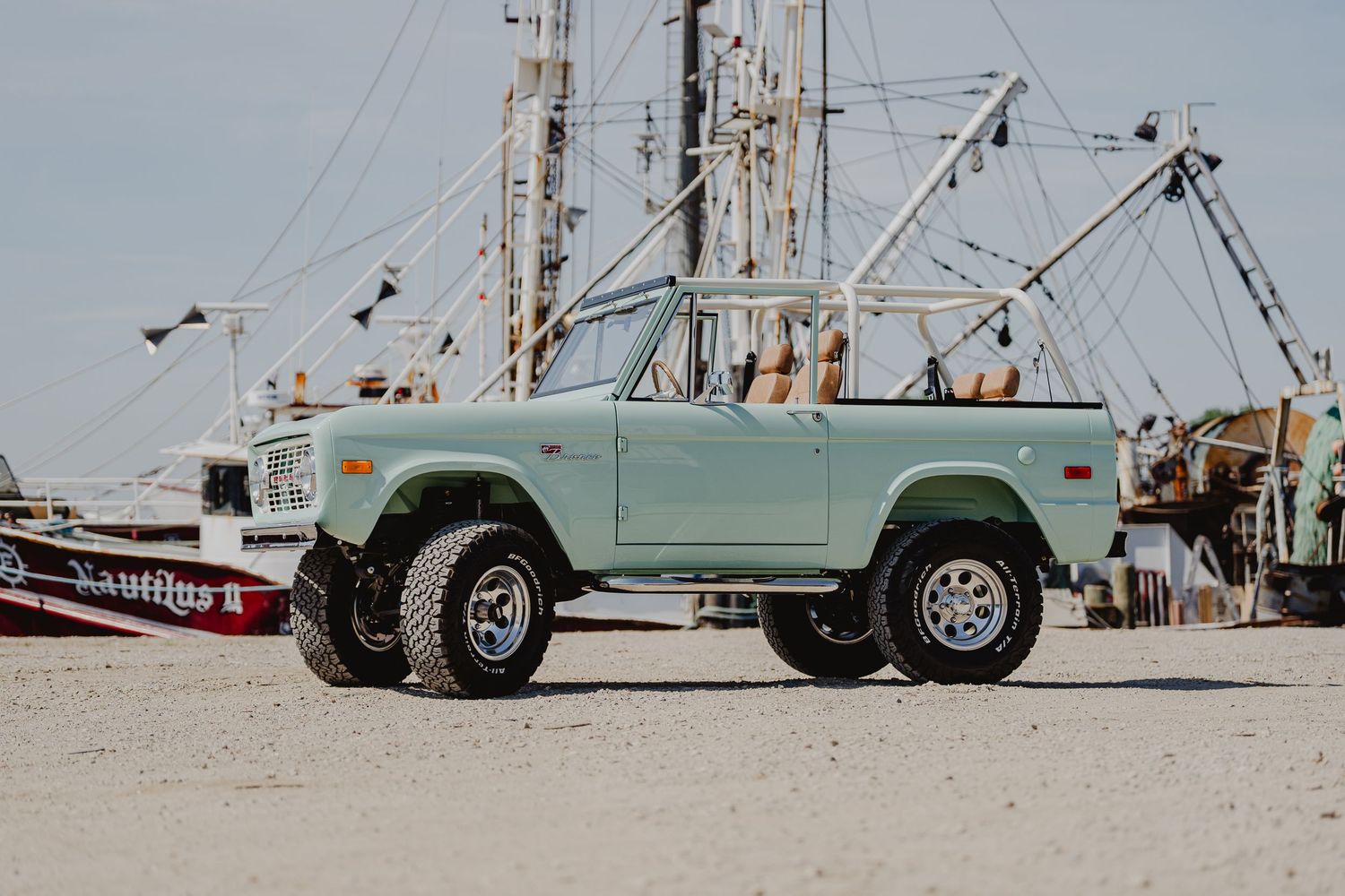 Vintage mint green Ford Bronco 4x4 lifted truck parked on sandy beach with shrimp boats in background.