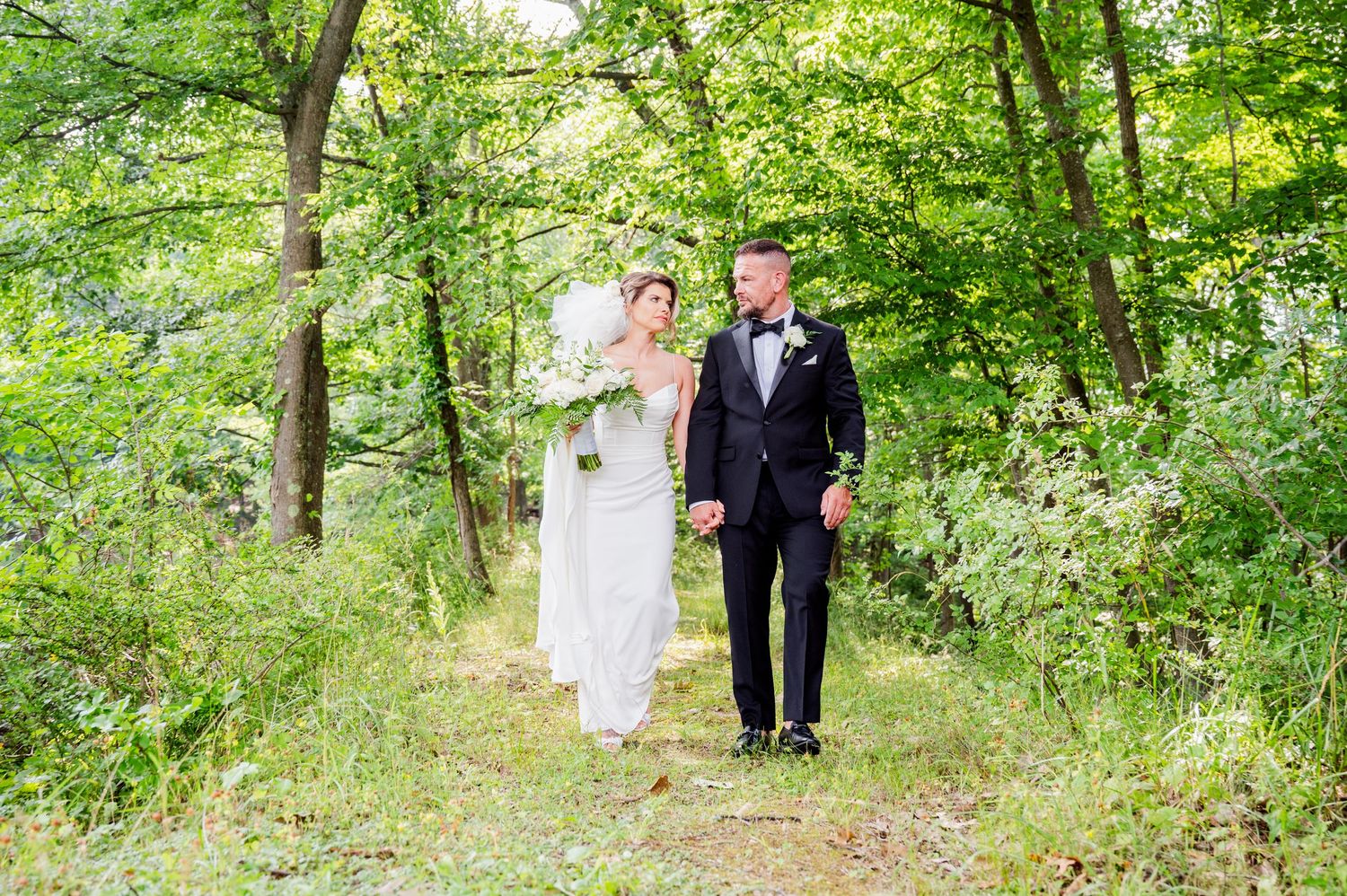 Wedding couple walking through a scenic wooded path with bright green foliage overhead.