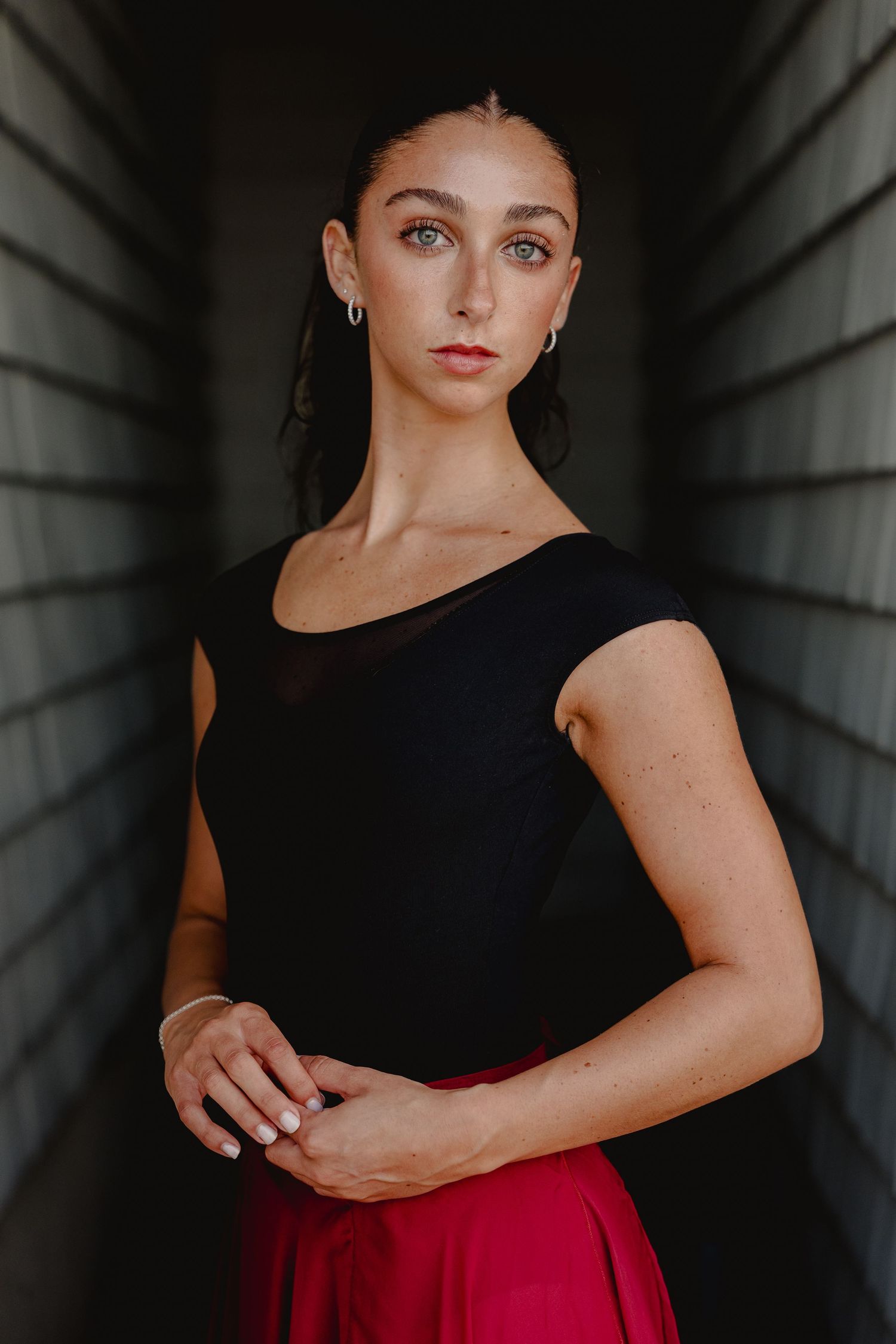 Young woman in black top and red skirt poses gracefully in a narrow corridor, looking directly at camera.