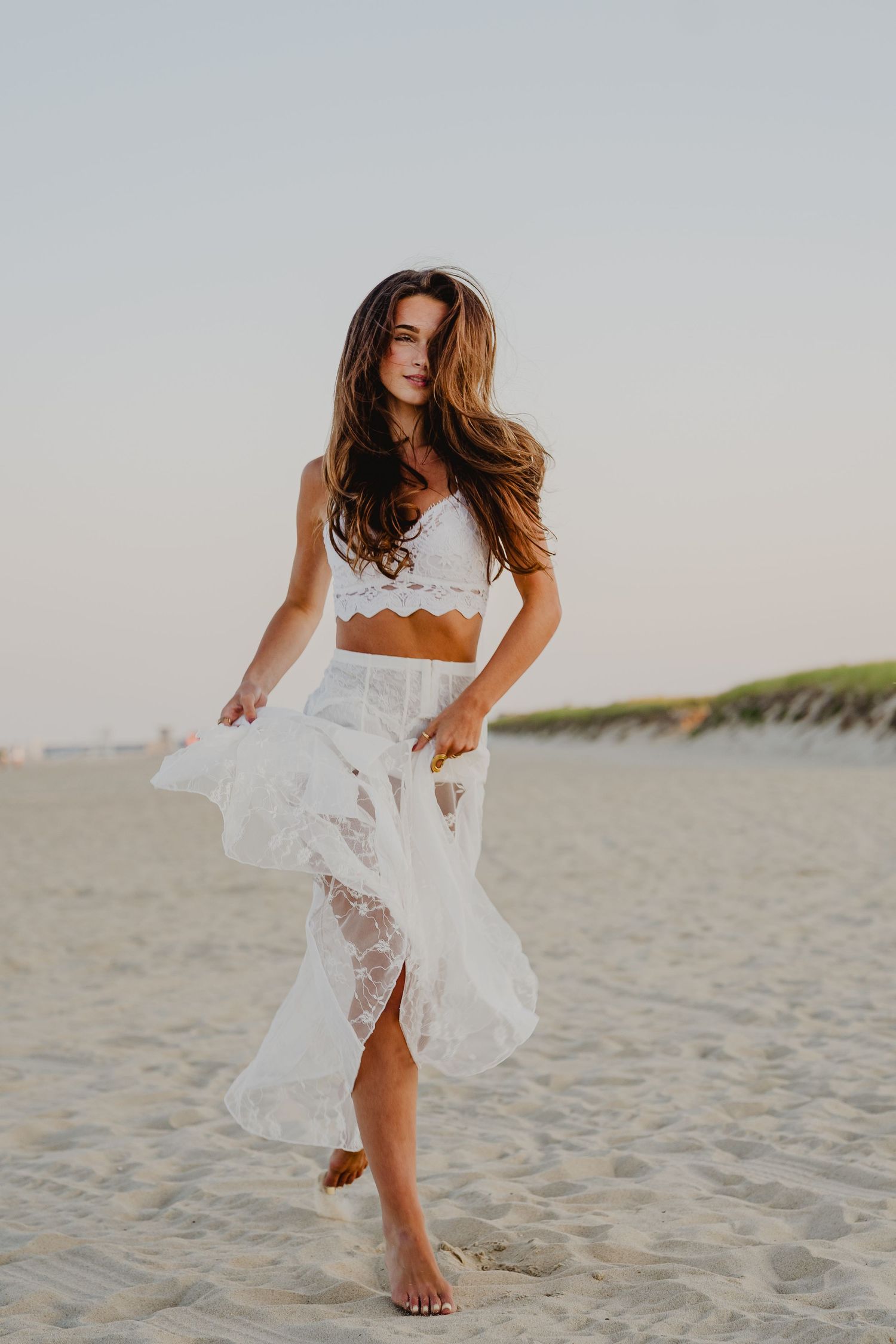 Brunette woman in white lace two-piece outfit walks on sandy beach at sunset.