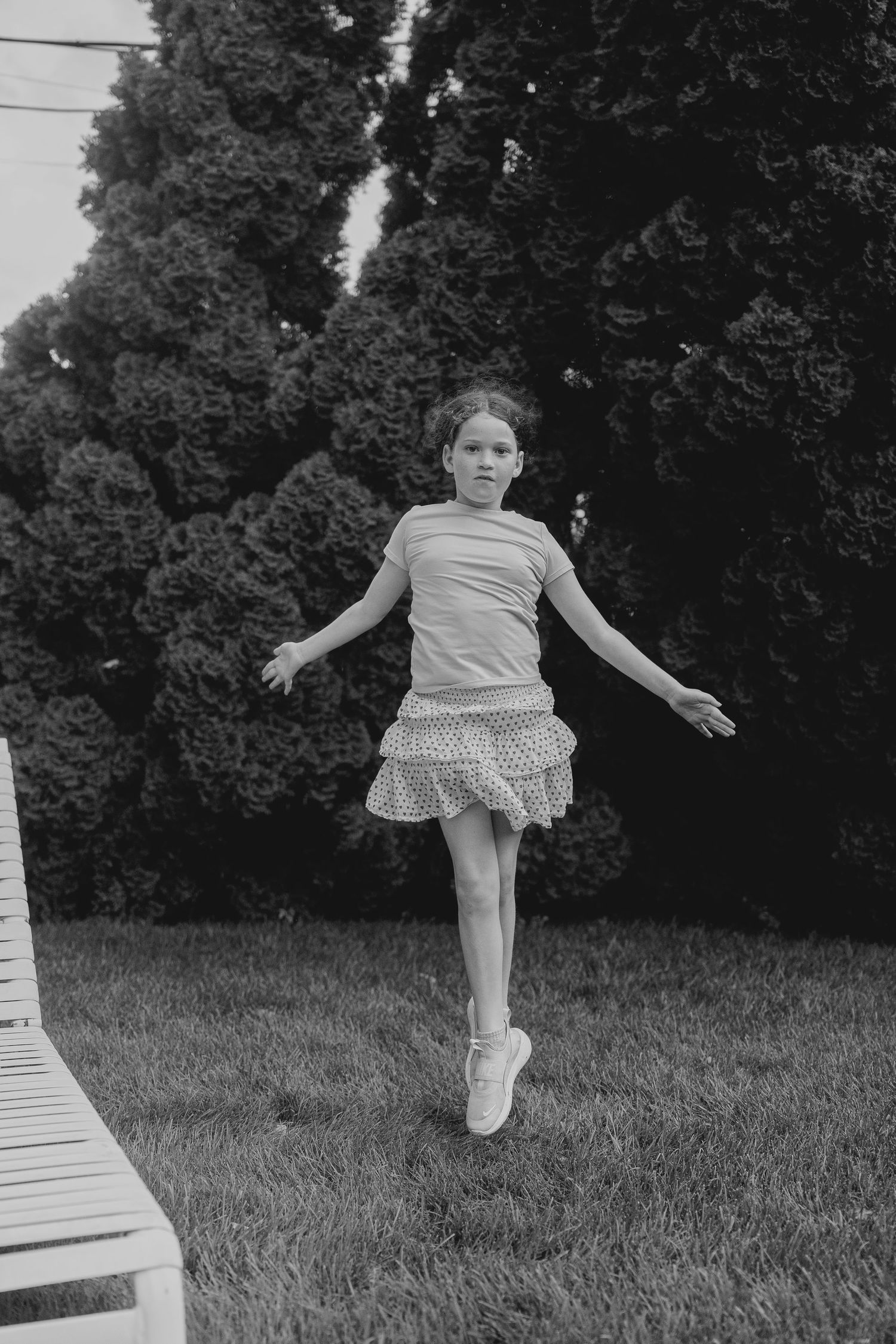 Black and white photo of a young girl in a skirt jumping outdoors with trees in the background.