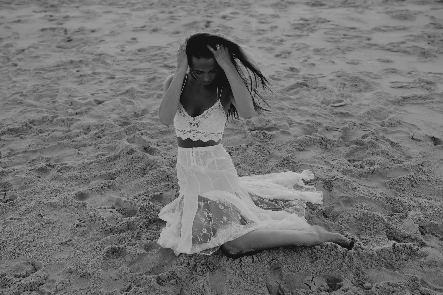 Black and white photo of a woman in a white dress sitting on sandy beach with hair blowing in the wind.