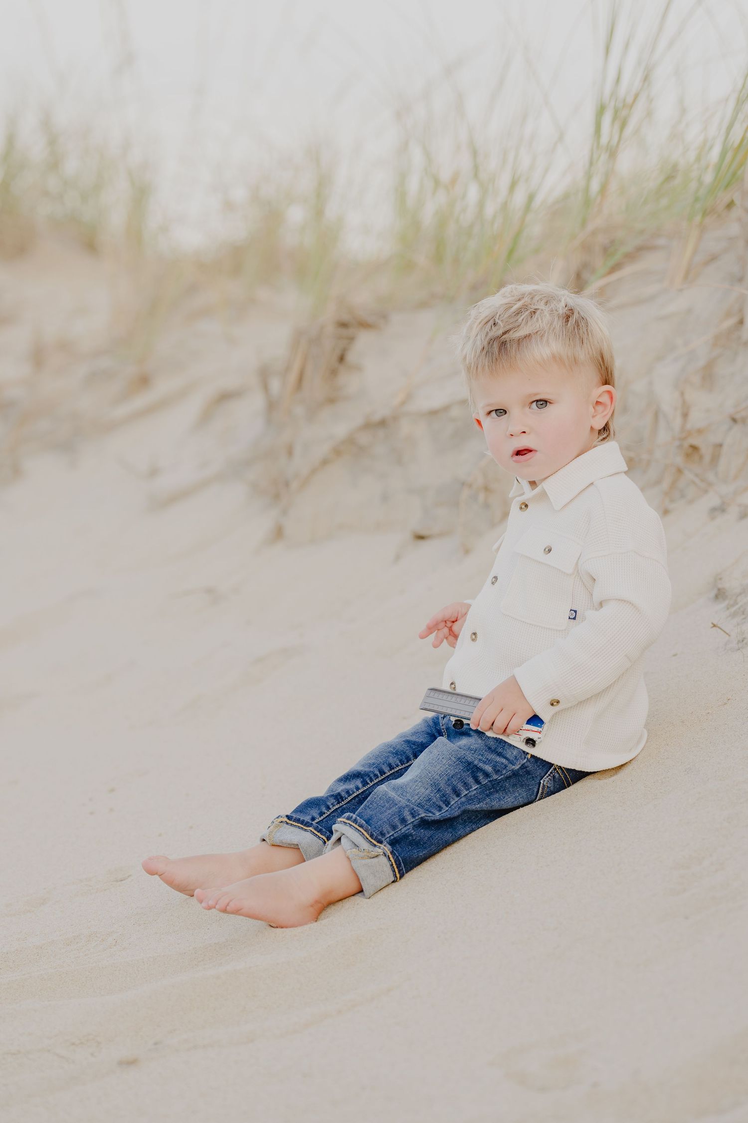 Toddler boy in white shirt and jeans sitting barefoot on sandy beach dunes.