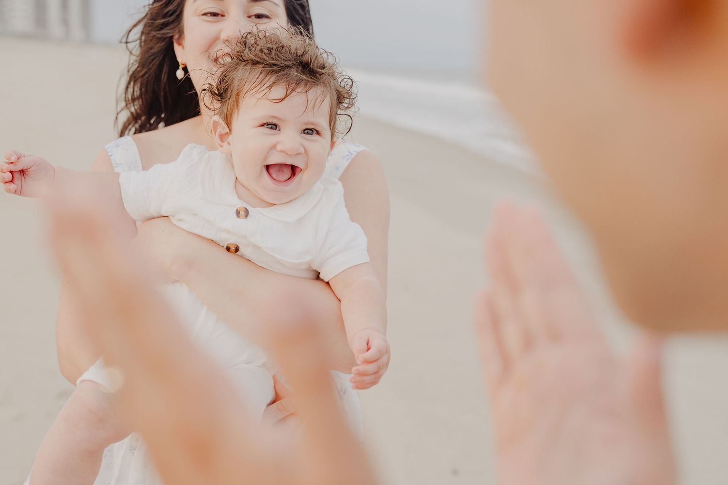 A joyful baby in a white outfit laughs while being held by a woman on a bright sandy beach.