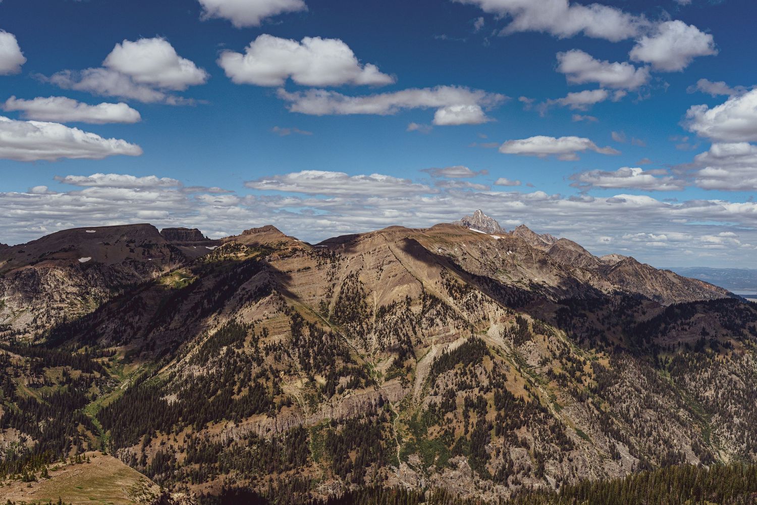 Panoramic mountain landscape with rugged peaks and ridges under blue sky with scattered white clouds in wilderness terrain.