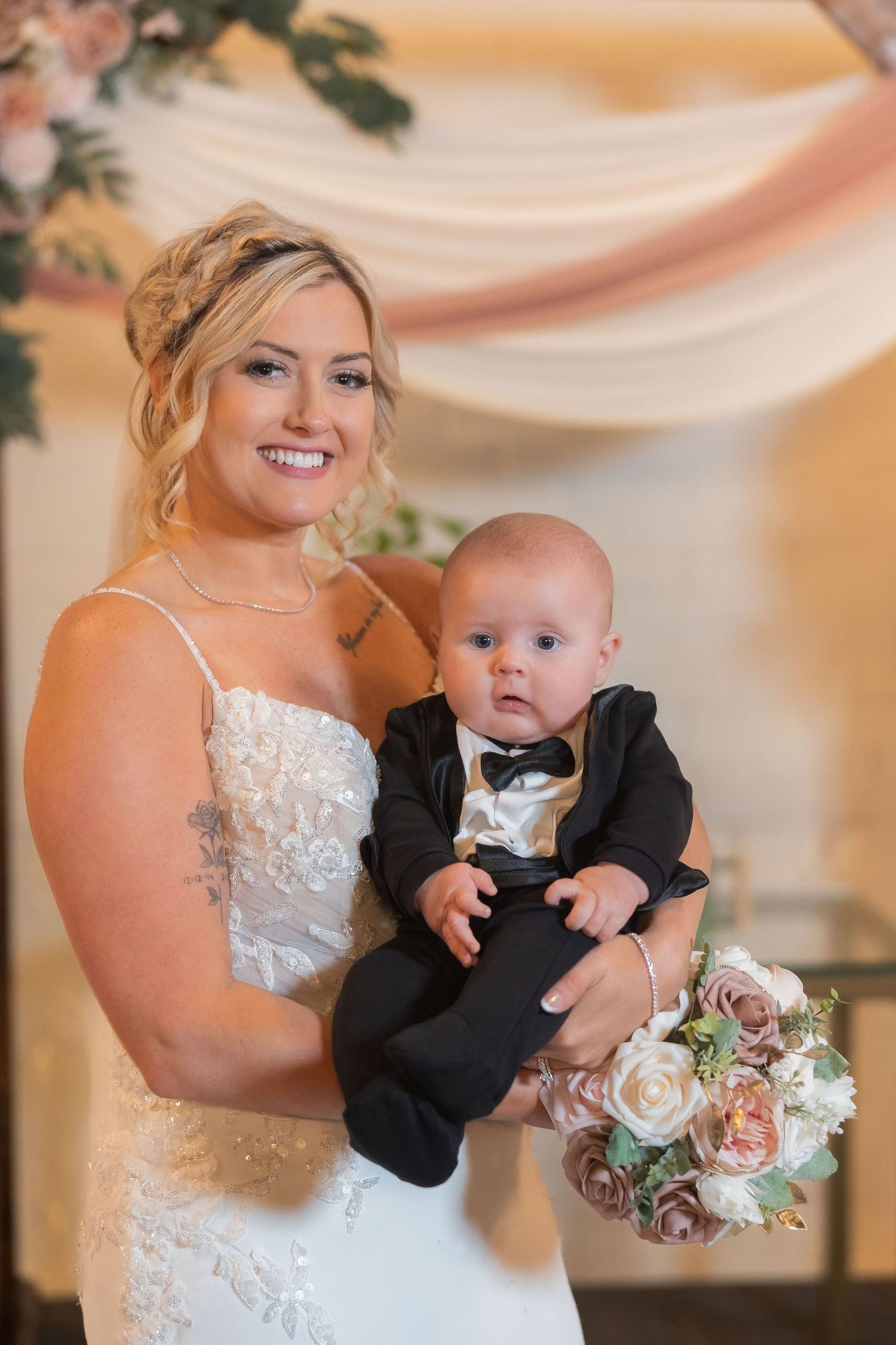 A bride in a white lace gown holds a baby wearing a tuxedo during a wedding photo session.