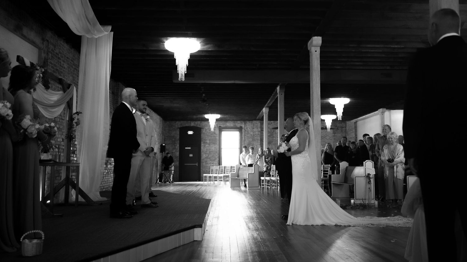 Dramatic black and white wedding ceremony photo with backlit silhouettes in an industrial venue.