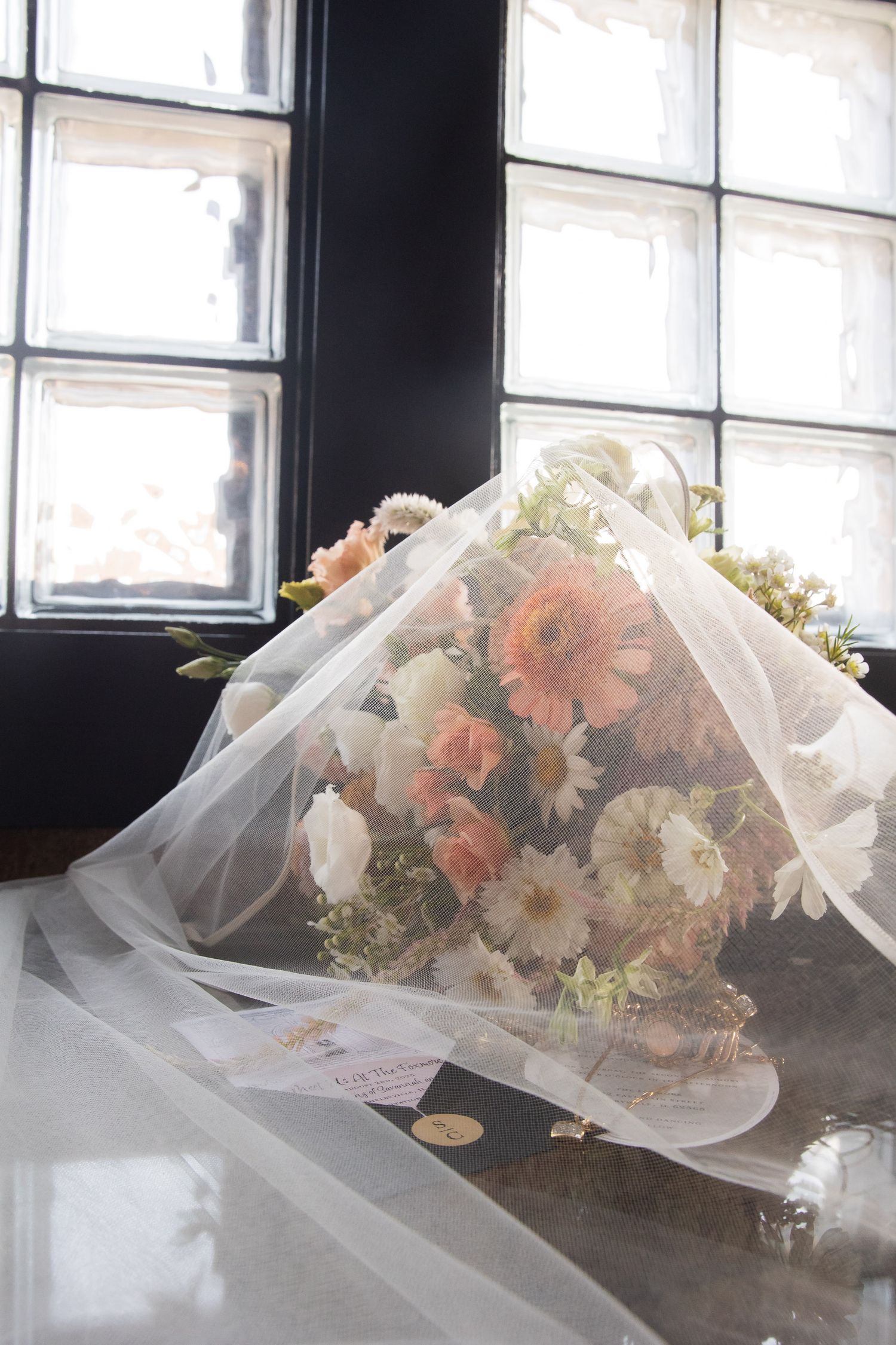 White and peach floral bouquet wrapped in delicate tulle veil against a window backdrop.