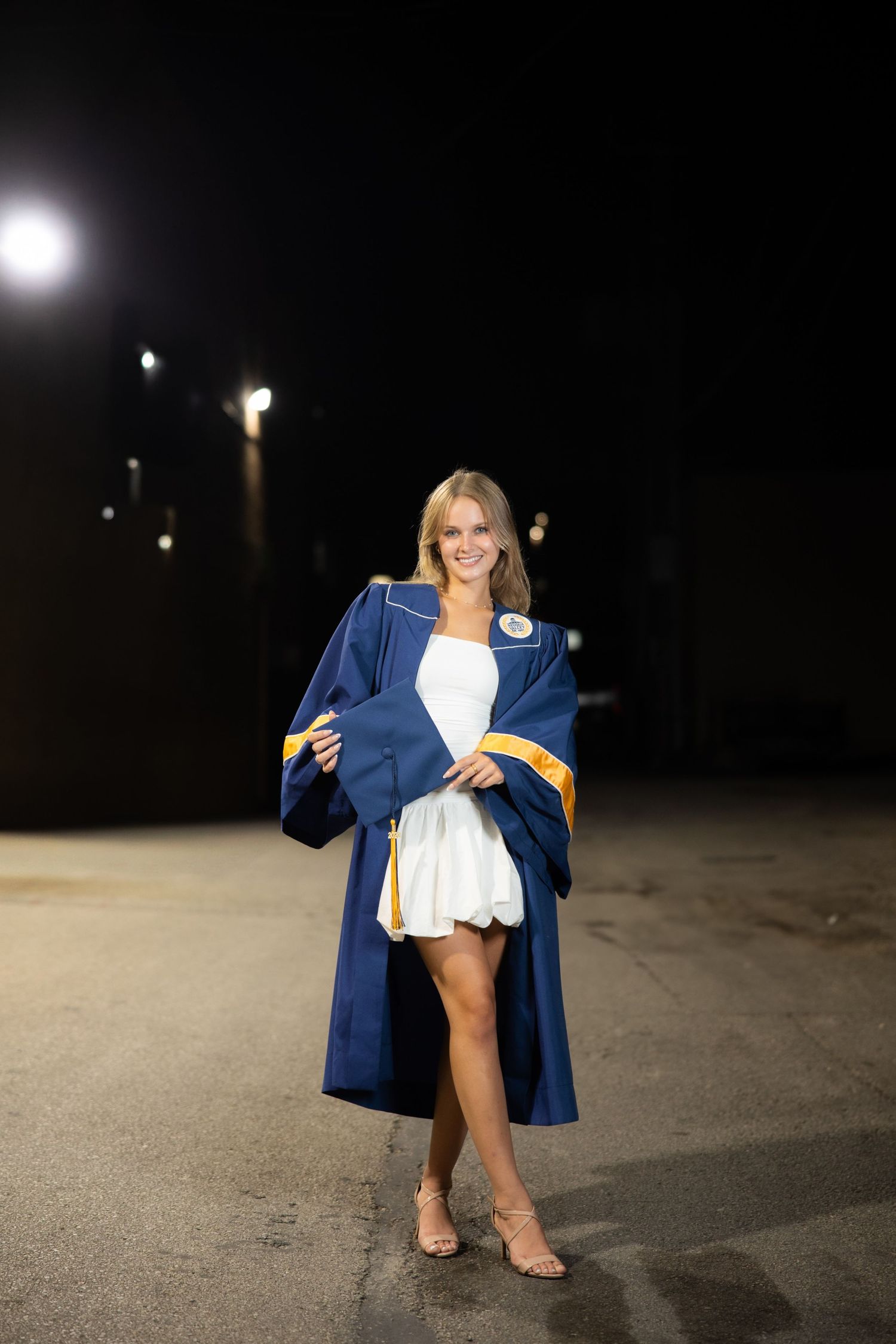A navy graduation gown captured in a nighttime photoshoot on pavement with ambient lighting.