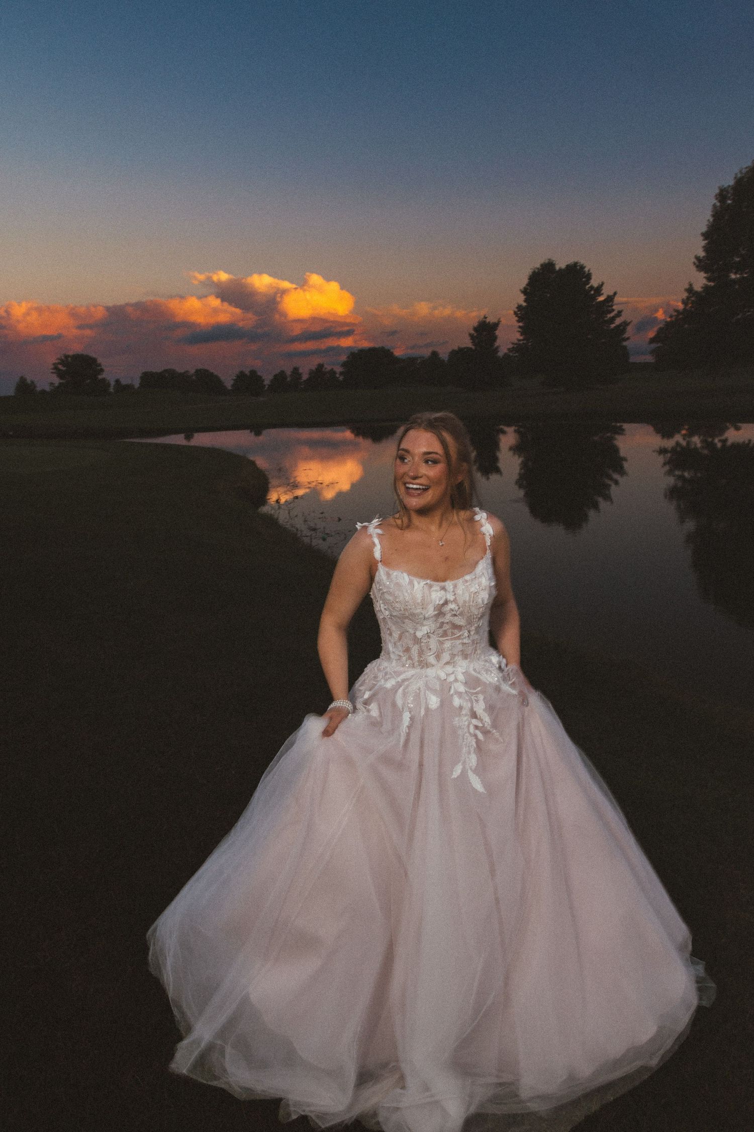Radiant bride in white lace ballgown smiles by a lake at golden sunset.