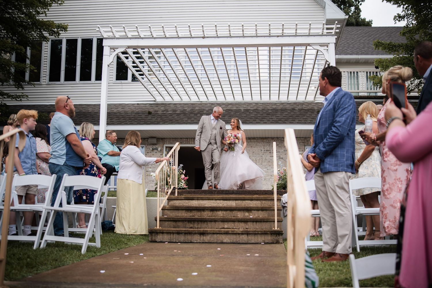 Bride walks down outdoor aisle with escort at elegant wedding ceremony with white chairs and pergola.