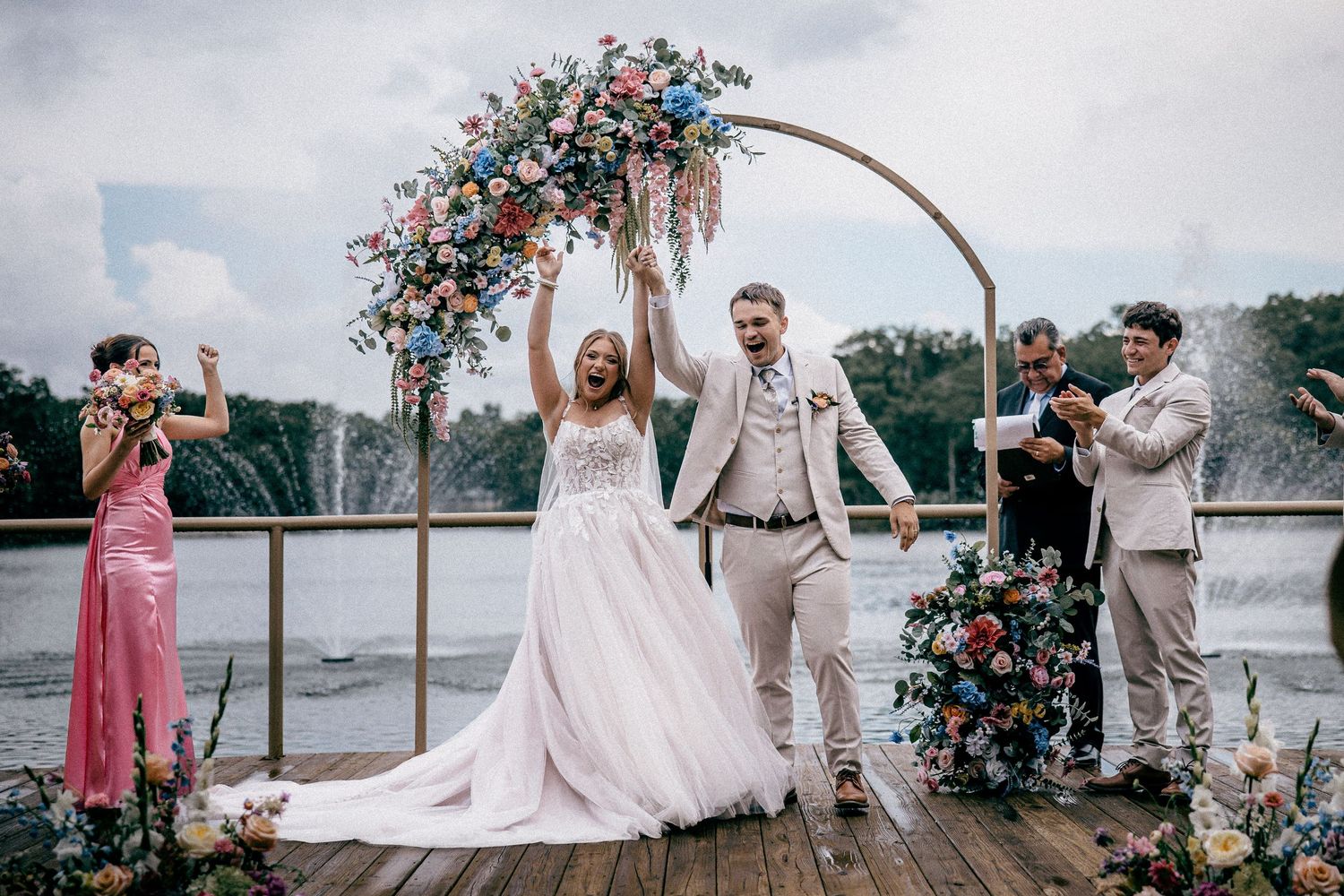 Joyful newlyweds celebrate under colorful floral arch on dock with fountain and guests cheering.