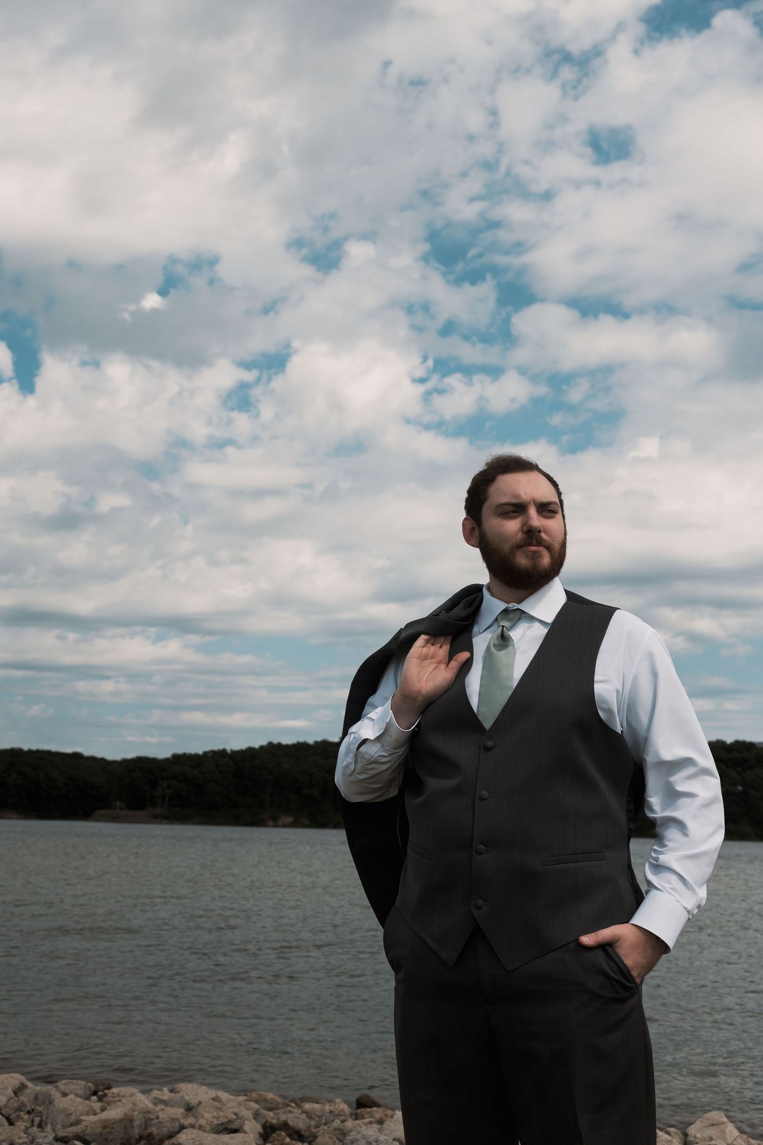 A well-dressed bearded man in a suit vest and tie poses by a rocky lakeside under a cloudy sky.