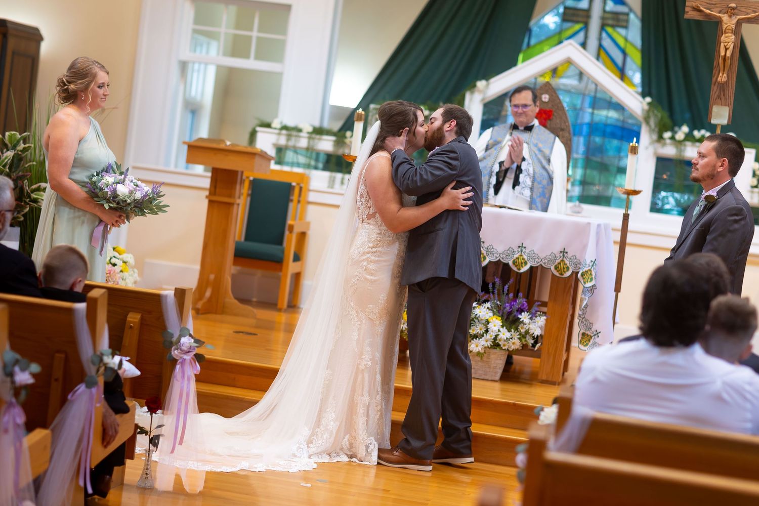 Bride and groom share first kiss at church wedding altar with officiant and bridesmaid standing nearby.