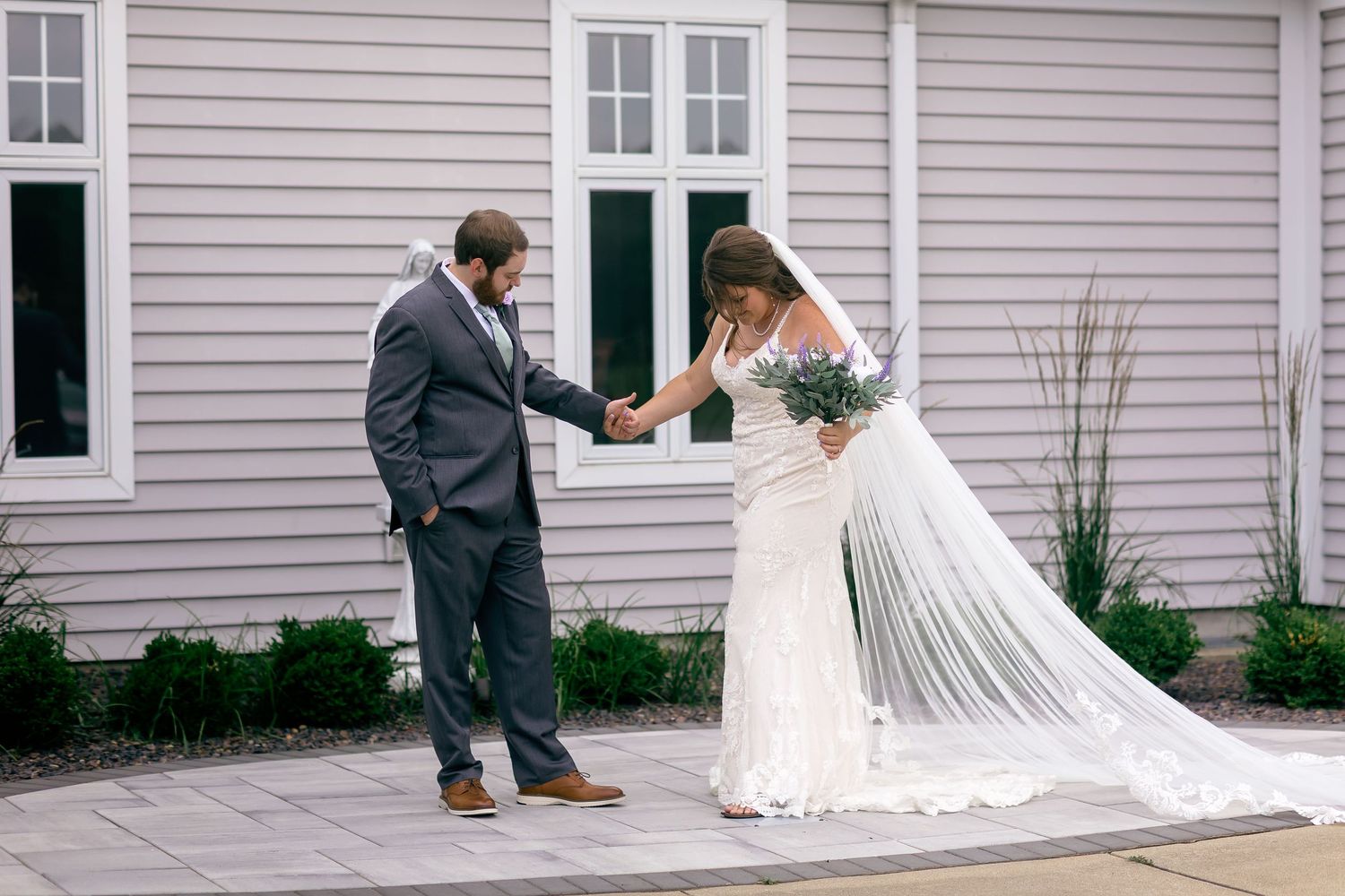 Groom in gray suit holds bride's hand as she curtsies in white lace wedding gown with veil outside gray building.