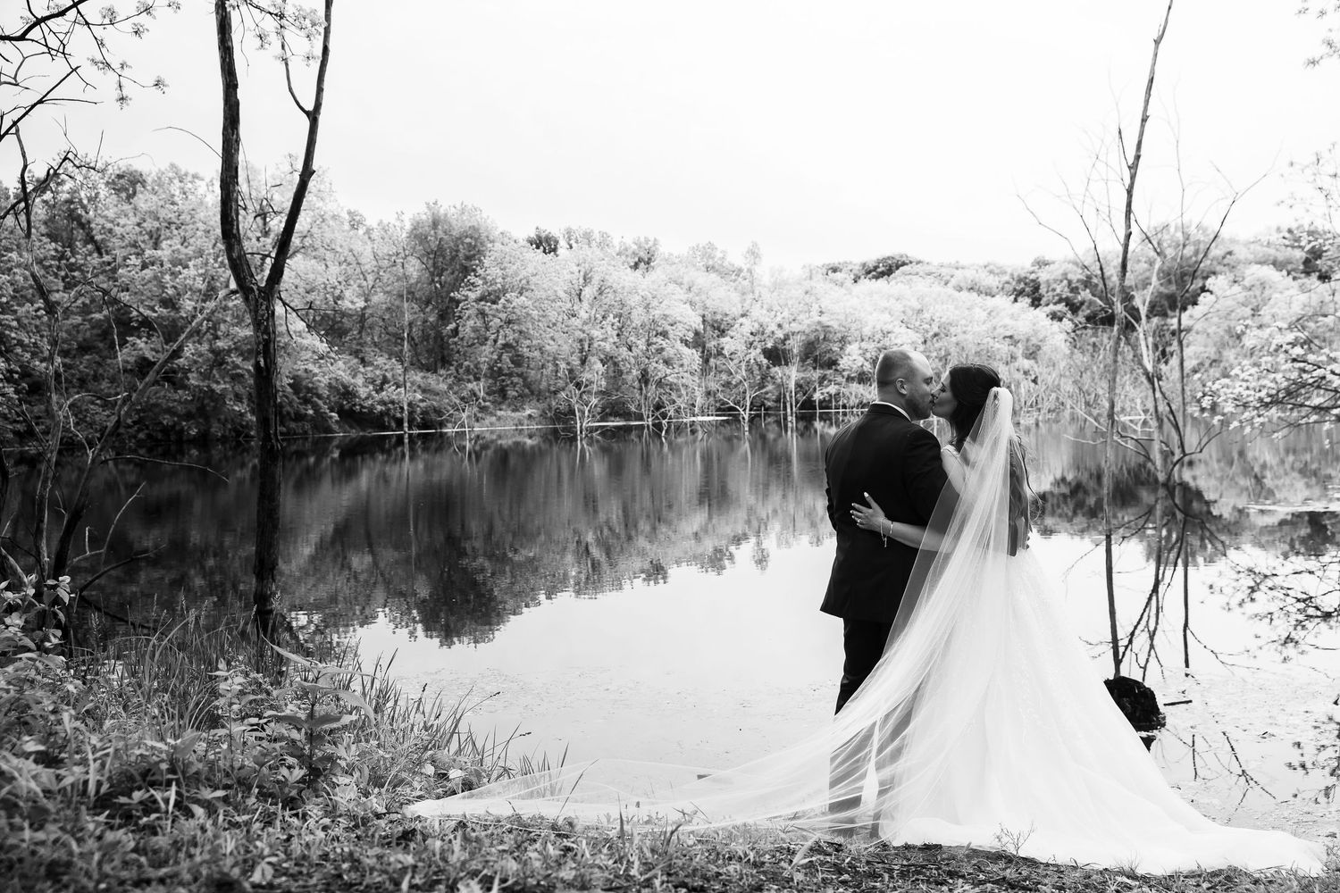 Black and white photo of bride and groom embracing beside a serene reflective lake after wedding.