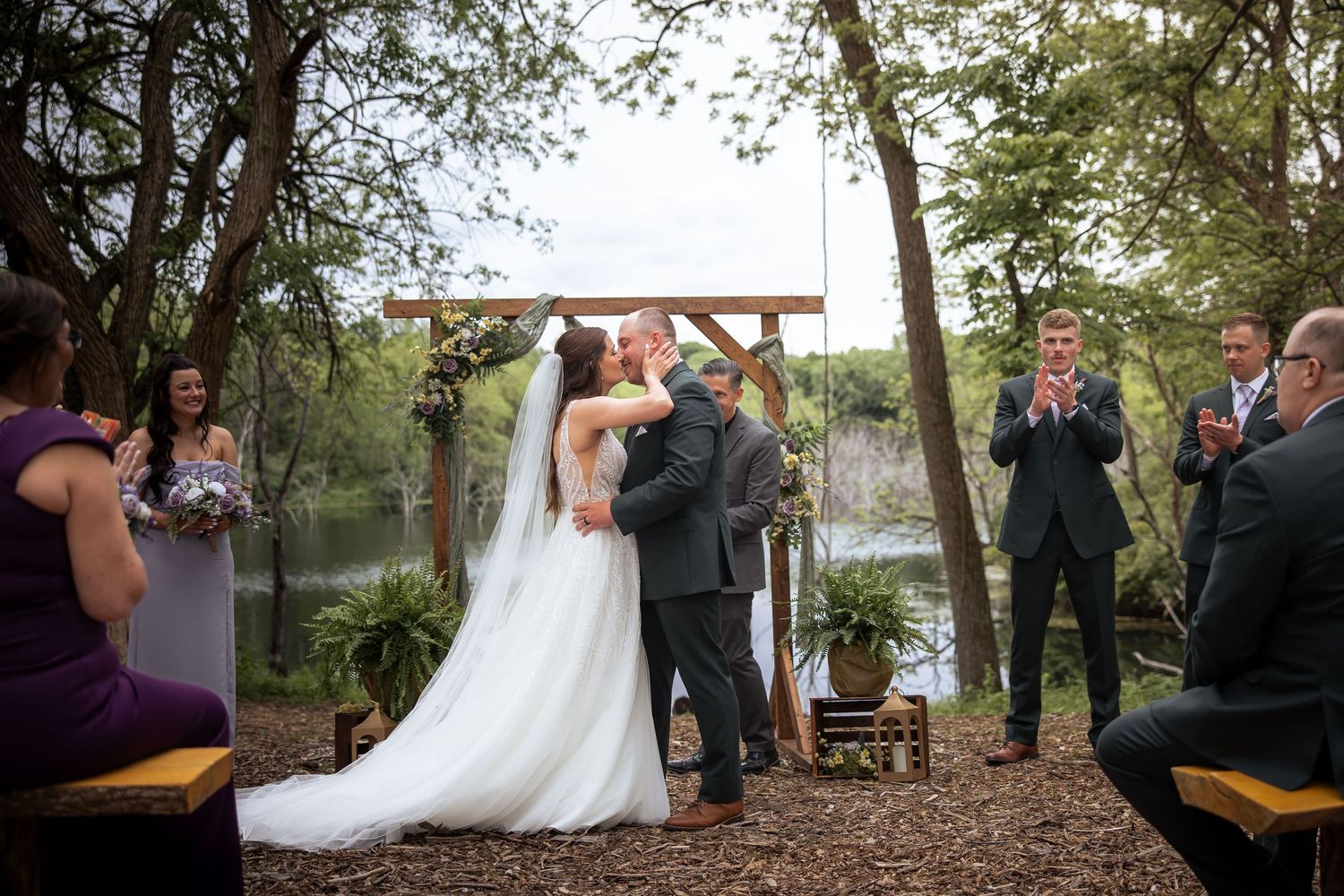 Bride and groom share first kiss at outdoor woodland wedding ceremony beside a serene lake.