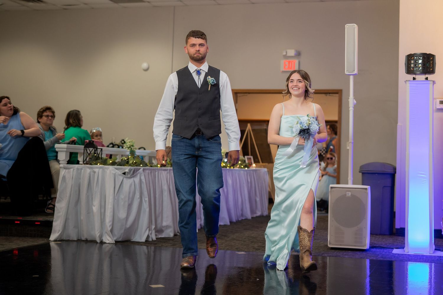 A couple walks into a wedding reception hall, man in vest and jeans, woman in mint green gown.