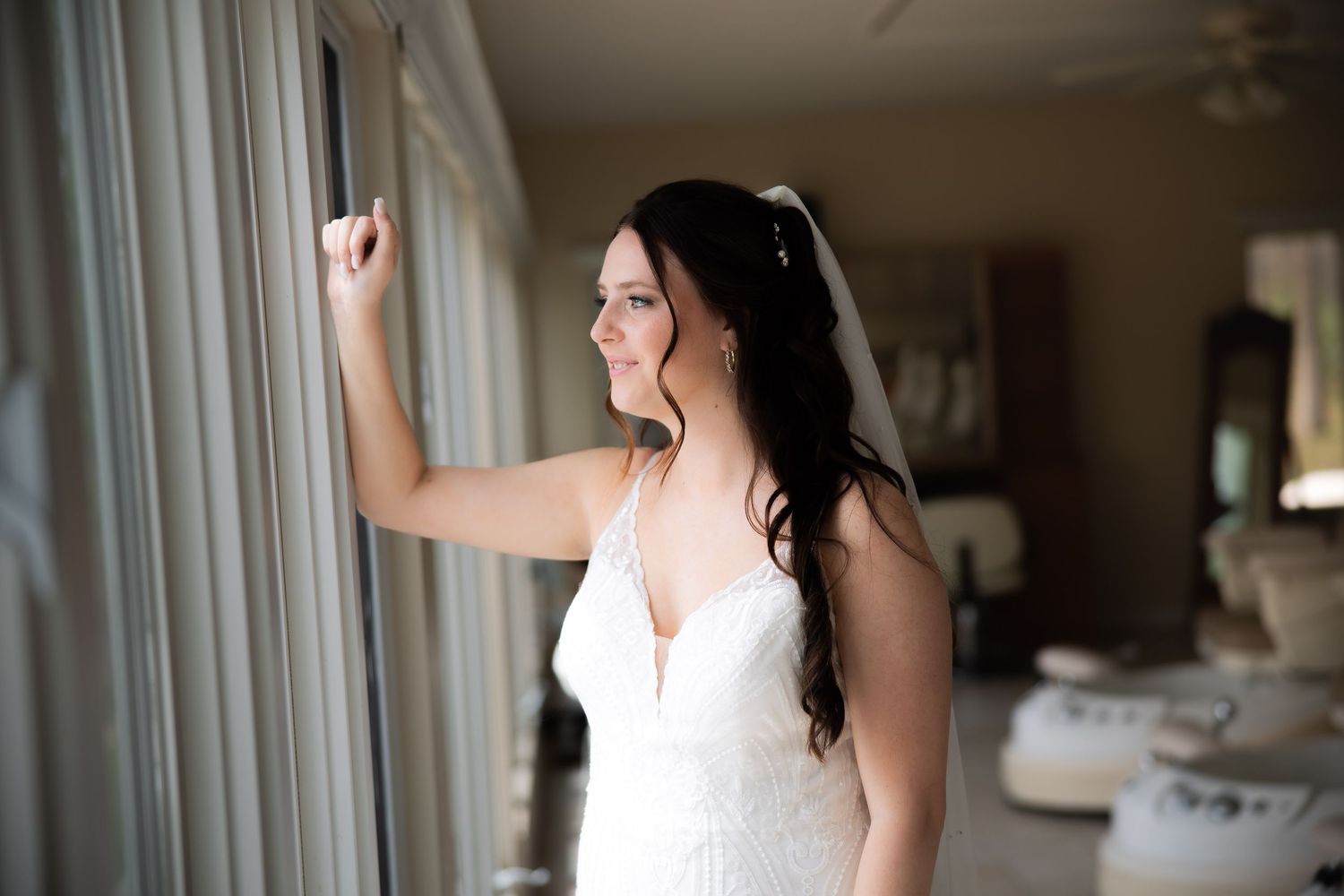 Bride in white lace wedding dress standing by window with soft natural light, long dark hair with veil.