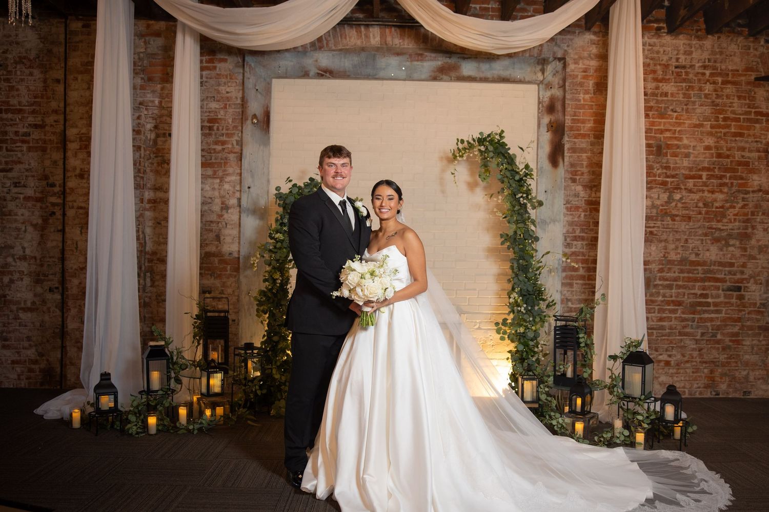 A bride and groom share an intimate moment in a rustic brick-walled venue with elegant white drapery and greenery.