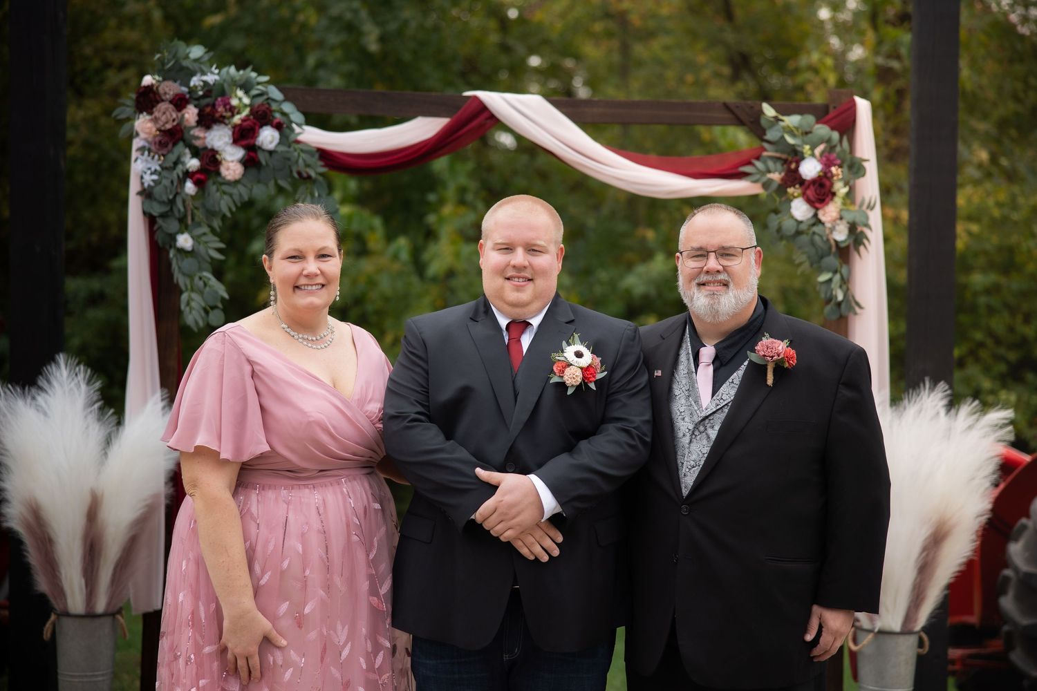 Wedding party poses together under white arch decorated with burgundy flowers in outdoor garden setting.